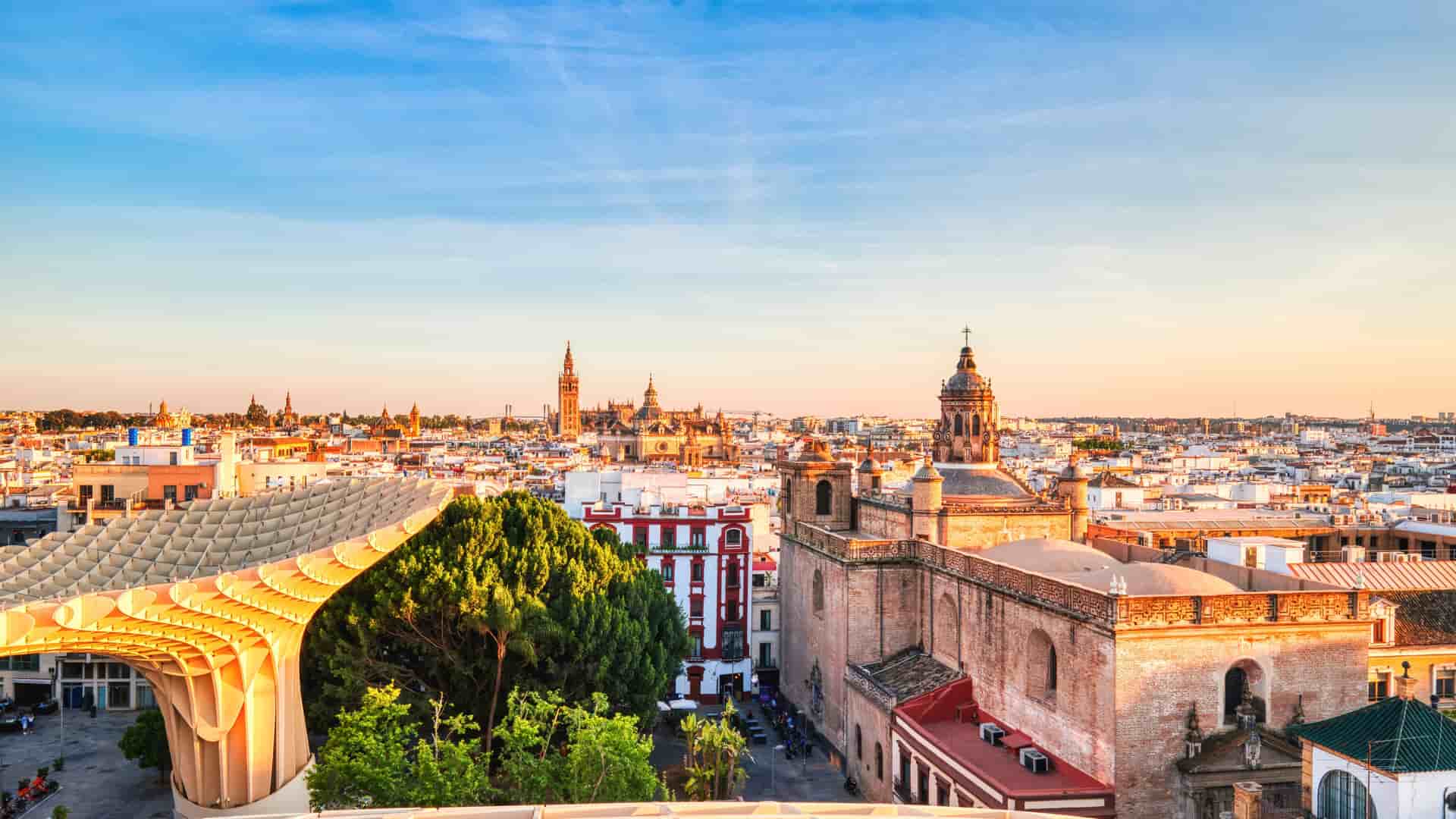 A scenic view of the iconic Plaza de España in Seville, Spain, featuring its grand arcade of arches and columns, a main building with a clock tower, and a bright blue sky.