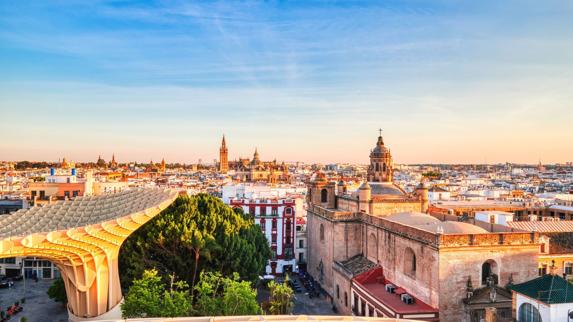 A scenic view of the iconic Plaza de España in Seville, Spain, featuring its grand arcade of arches and columns, a main building with a clock tower, and a bright blue sky.