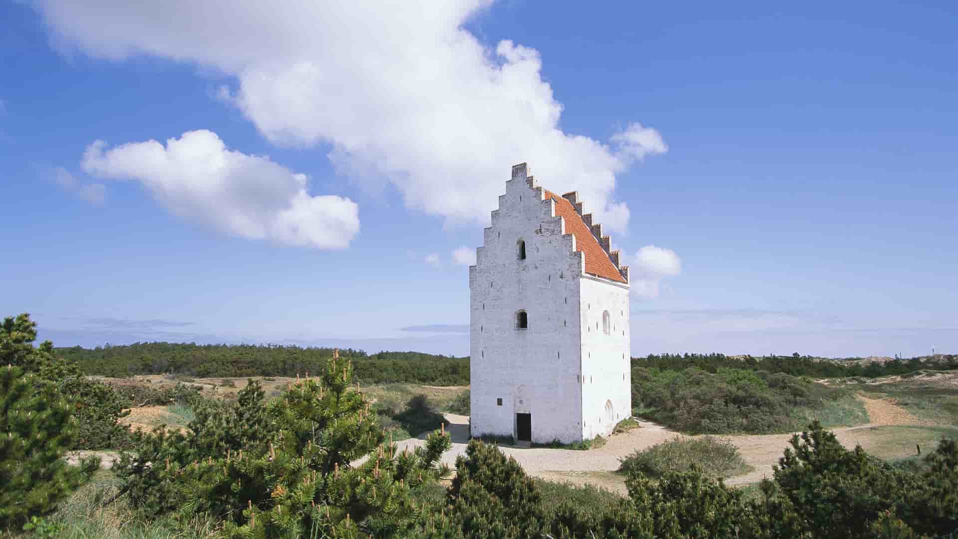 A striking image of Den Tilsandede Kirke, or the Buried Church, a historic white church with a red roof partially submerged in sand dunes near Skagen, Denmark.