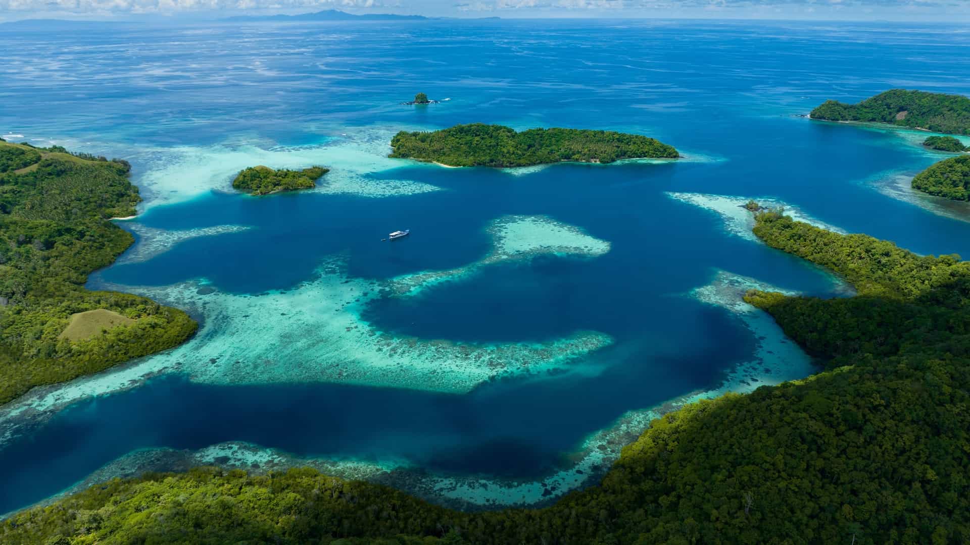 An aerial view of lush, forested tropical islands surrounding a deep blue bay with vibrant turquoise coral reefs and a single boat.