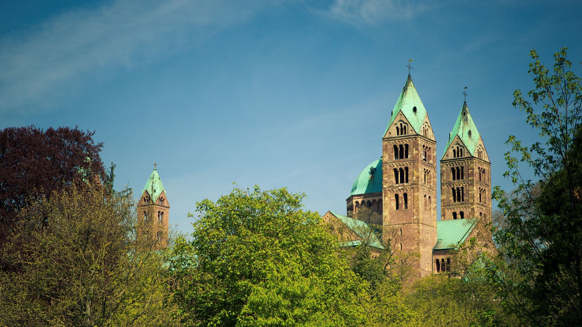A beautiful view of the Speyer Cathedral with its iconic twin green-roofed towers peeking above the lush green trees against a clear blue sky in Speyer, Germany.