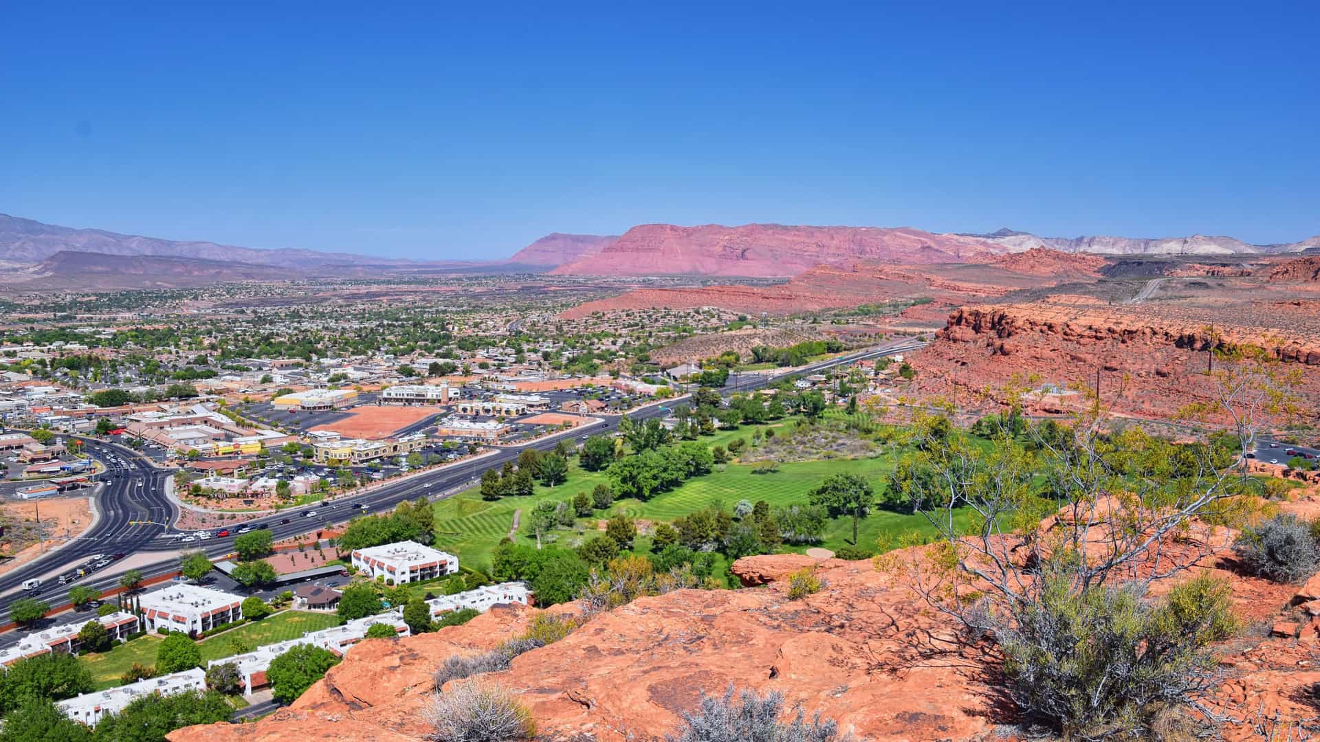 "A high-angle view of the city of St. George, Utah, with lush green golf courses and residential areas, surrounded by the dramatic red rock desert landscape and distant mountains under a clear sky. "