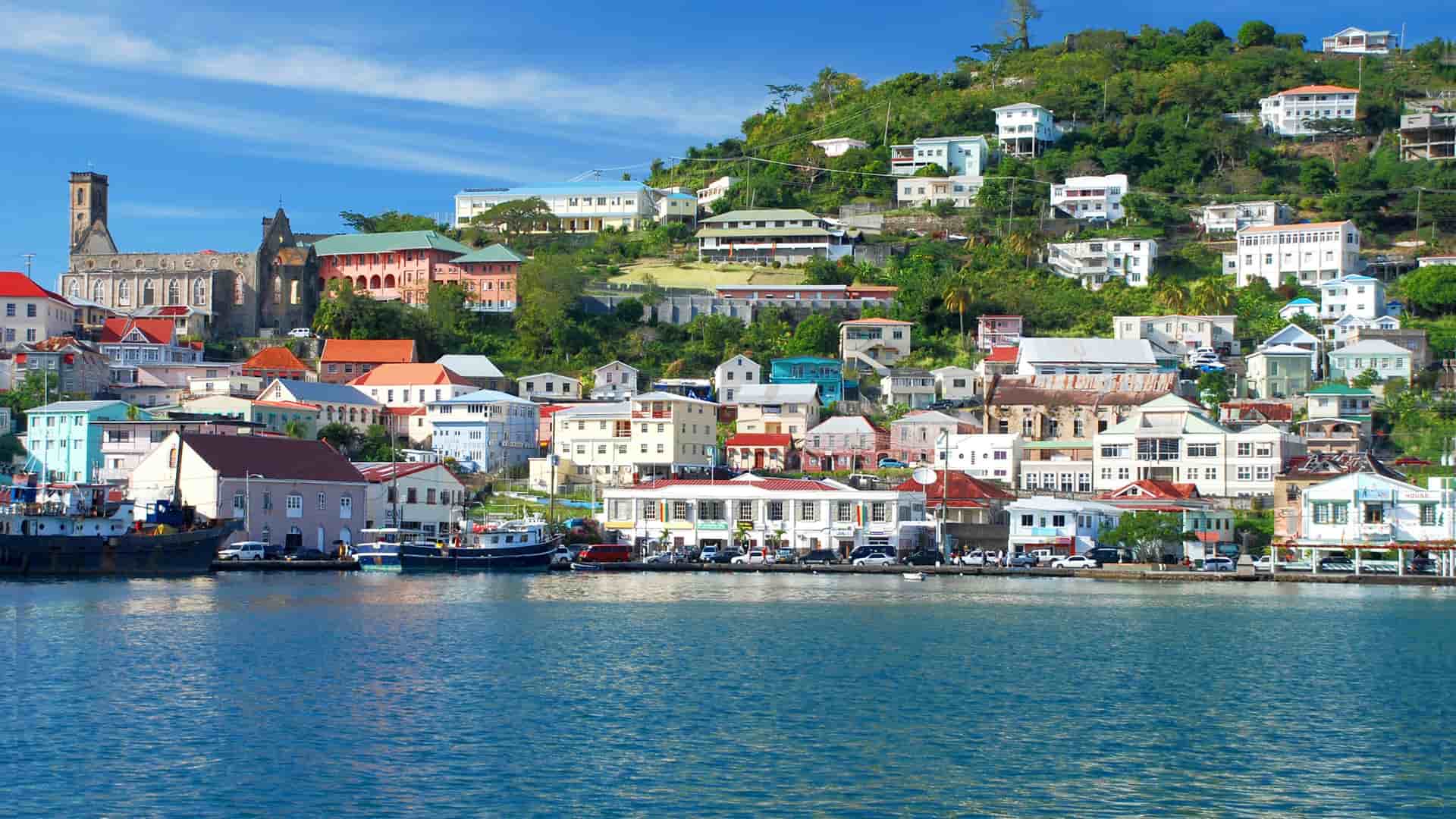 An aerial view of the colorful waterfront of St. George's, Grenada, with the town's buildings nestled on a hillside and a long, rocky breakwater extending into the calm, turquoise water.