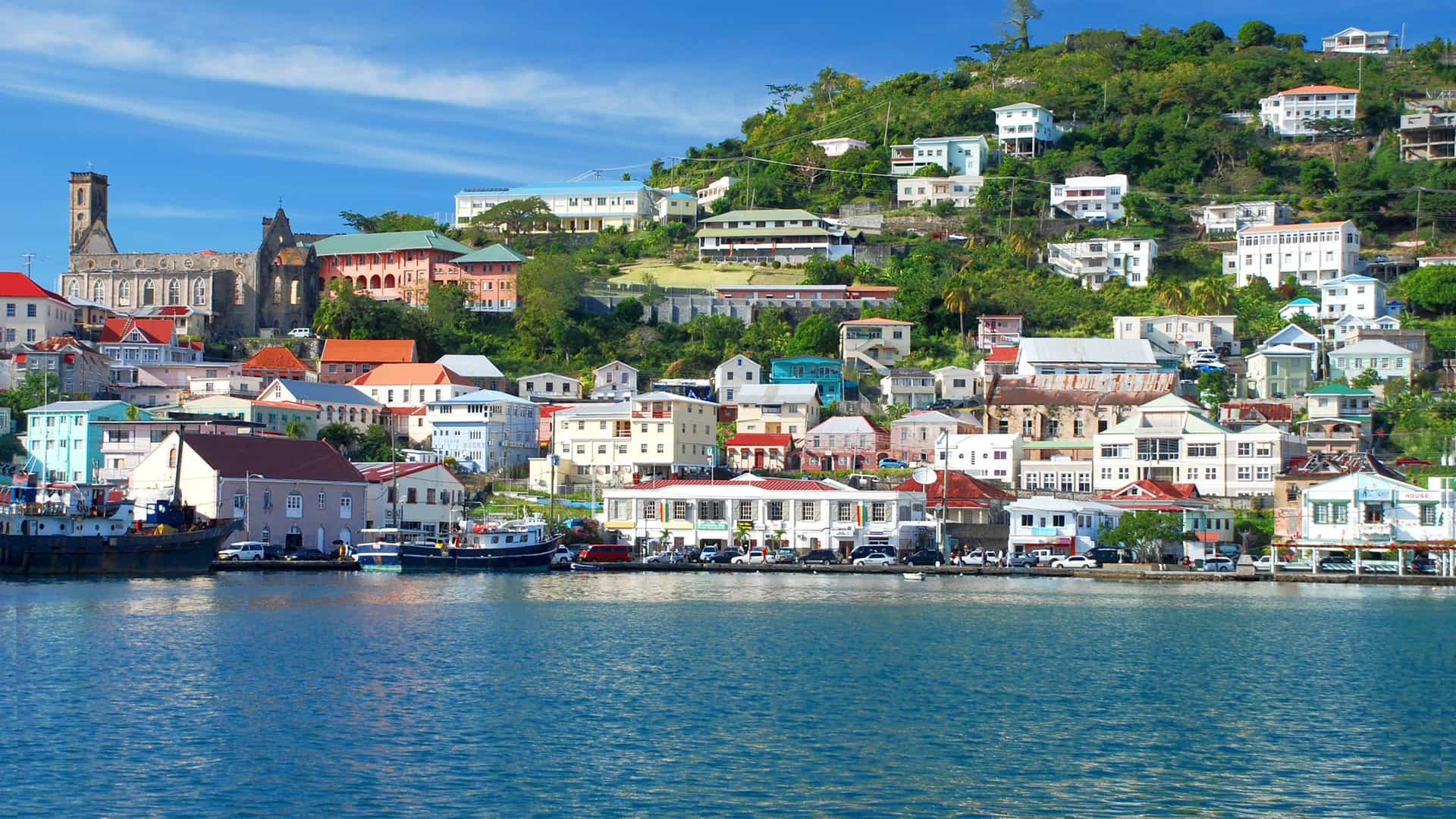 An aerial view of the colorful waterfront of St. George's, Grenada, with the town's buildings nestled on a hillside and a long, rocky breakwater extending into the calm, turquoise water.