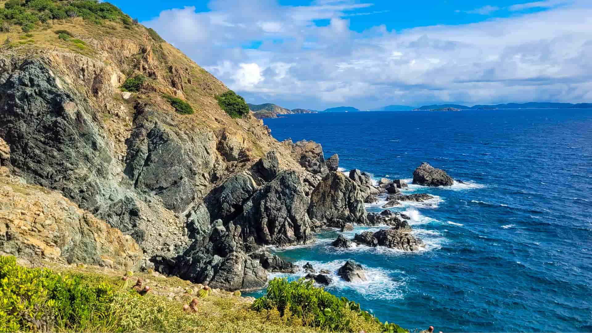A high-angle view of the rugged, rocky coastline of St. John, US Virgin Islands, with the deep blue sea crashing against the cliffs and islands in the distance under a cloudy blue sky.