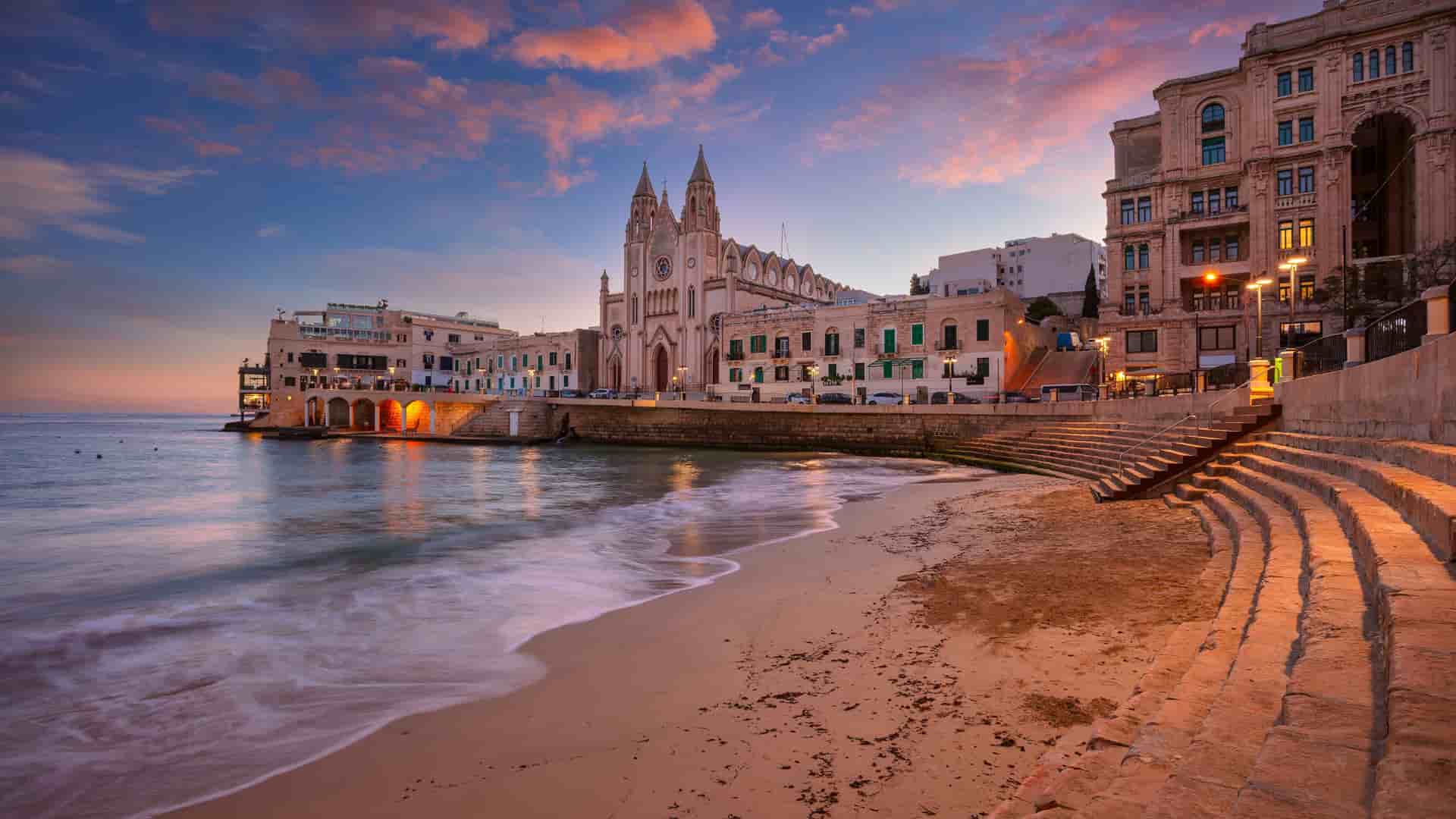 "A breathtaking sunset view of St. Julian's, Malta, with a sandy beach and gentle waves, showcasing the iconic Parish Church of Our Lady of Mount Carmel and the illuminated town buildings in the background. "