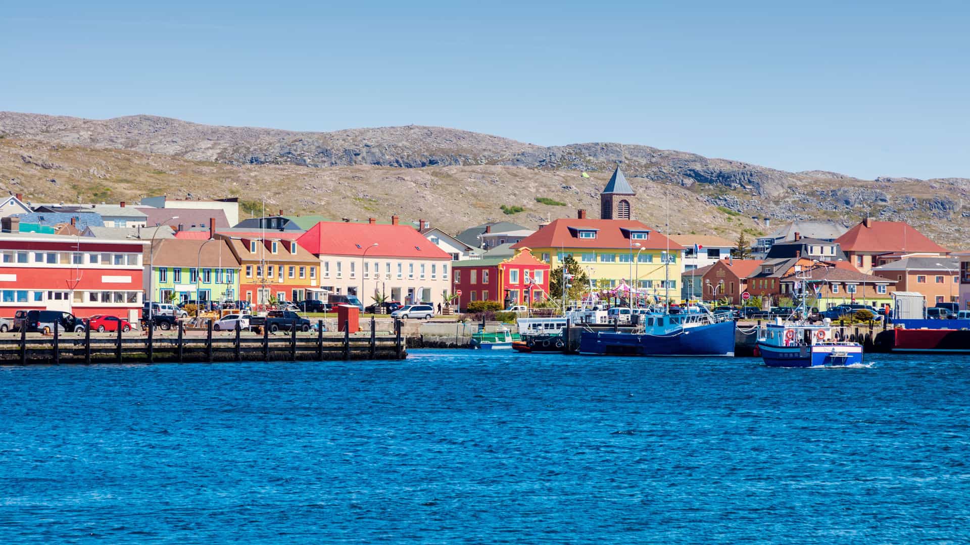 A vibrant view of the colorful waterfront of Saint-Pierre, Saint Pierre and Miquelon, with its French-style buildings and boats docked at the harbor in front of a rocky, barren landscape.