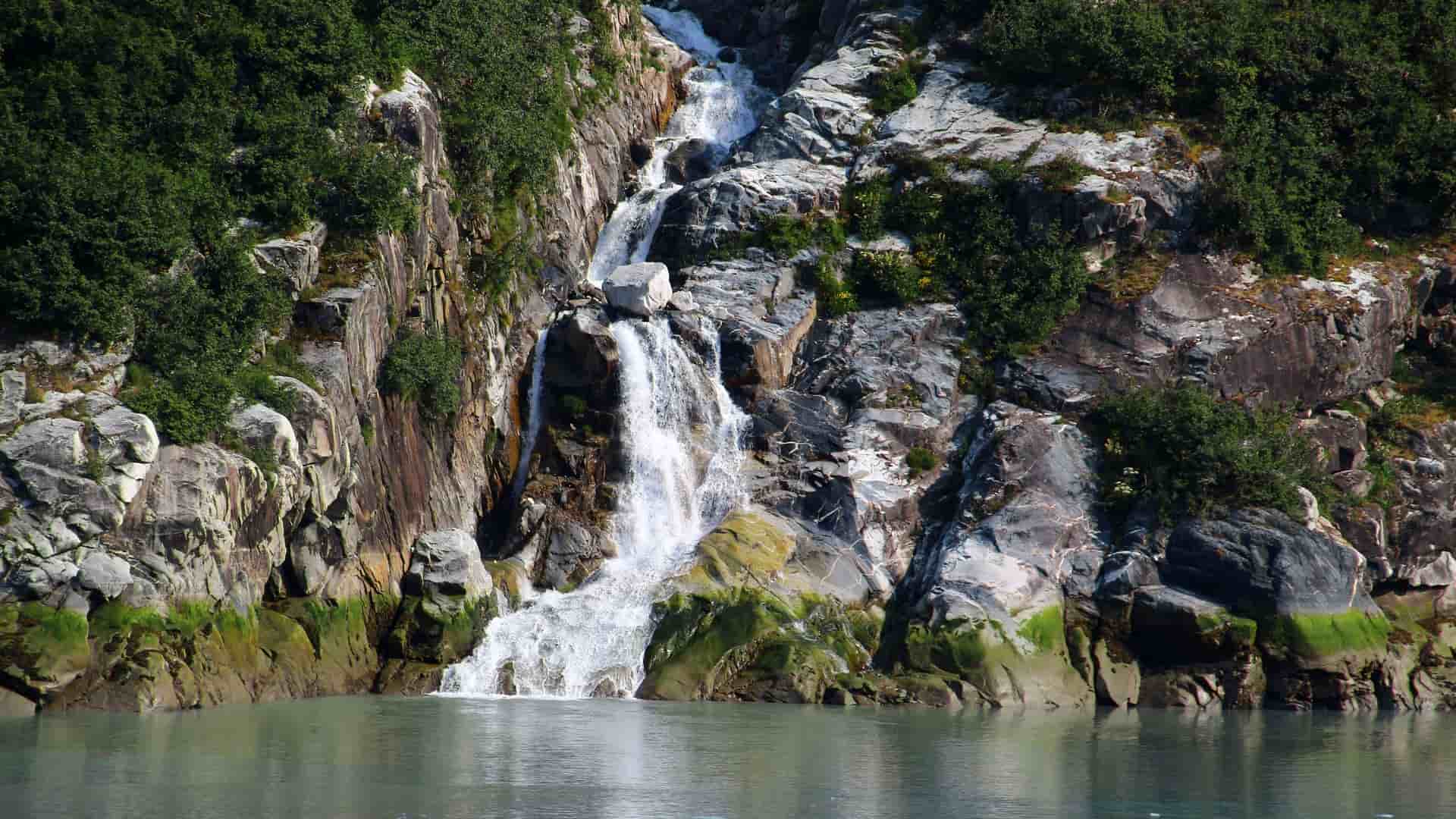 A wide-angle view of a cascading waterfall flowing down a steep, rocky cliff into the calm, gray waters of Stephens Passage, Alaska, surrounded by lush green foliage.
