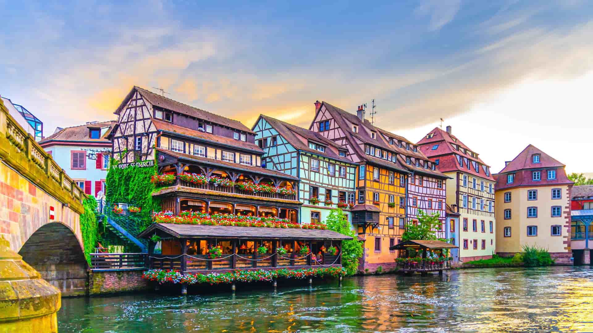 A wide-angle view of the colorful half-timbered buildings in Petite France, Strasbourg, with a bridge over the Ill River and flowers hanging from the balconies.