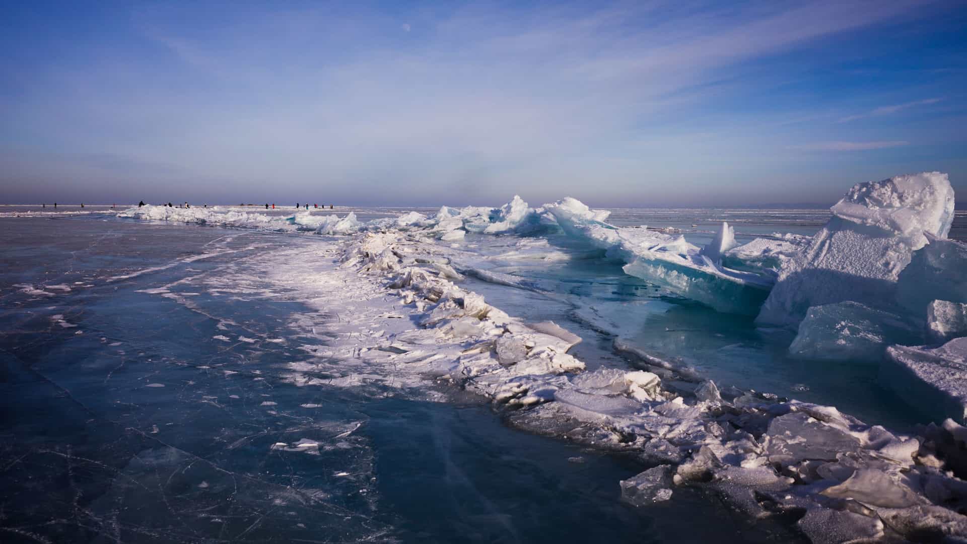 A frozen expanse in Strezelecki Harbour with cracked sheets of ice piled into jagged ridges across a vast icy surface, distant figures standing on the frozen landscape beneath a clear blue sky.