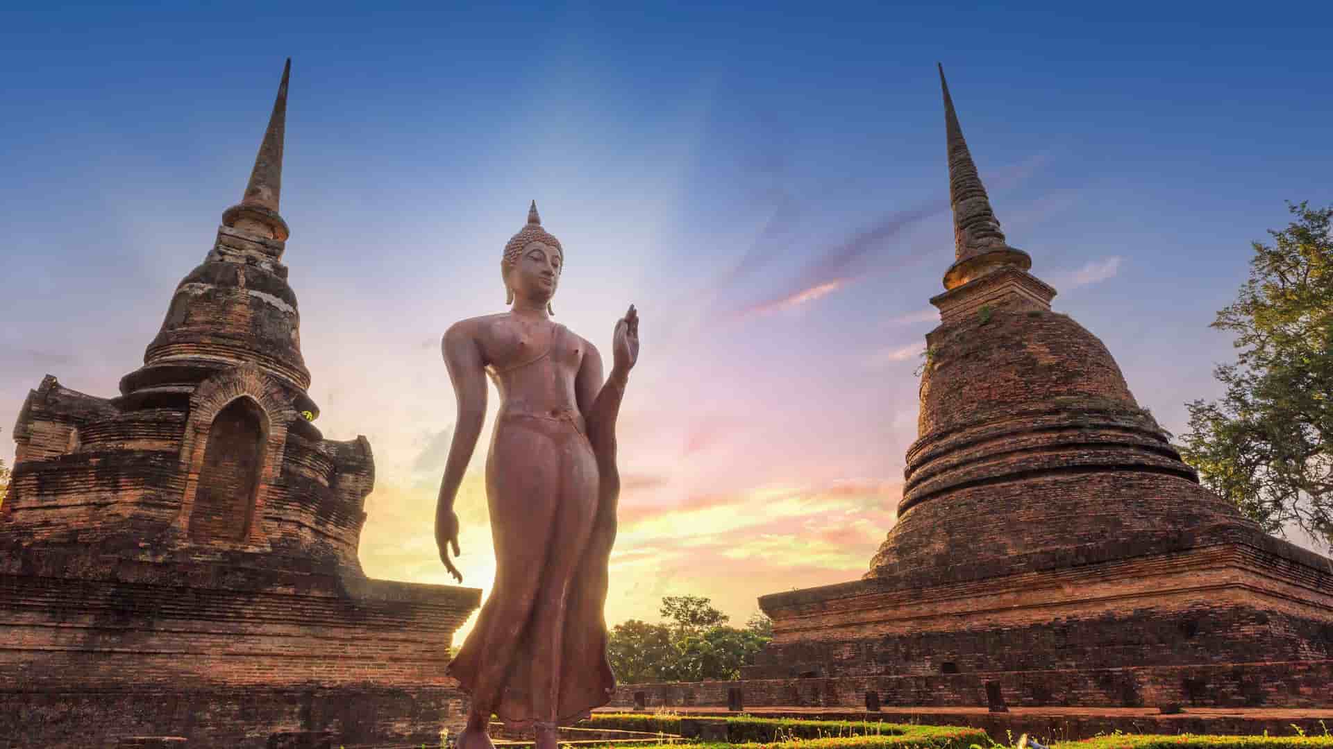 "A towering bronze Buddha statue with a distinctive walking pose, standing between two ancient brick temple ruins at the historic Sukhothai Historical Park in Thailand. "