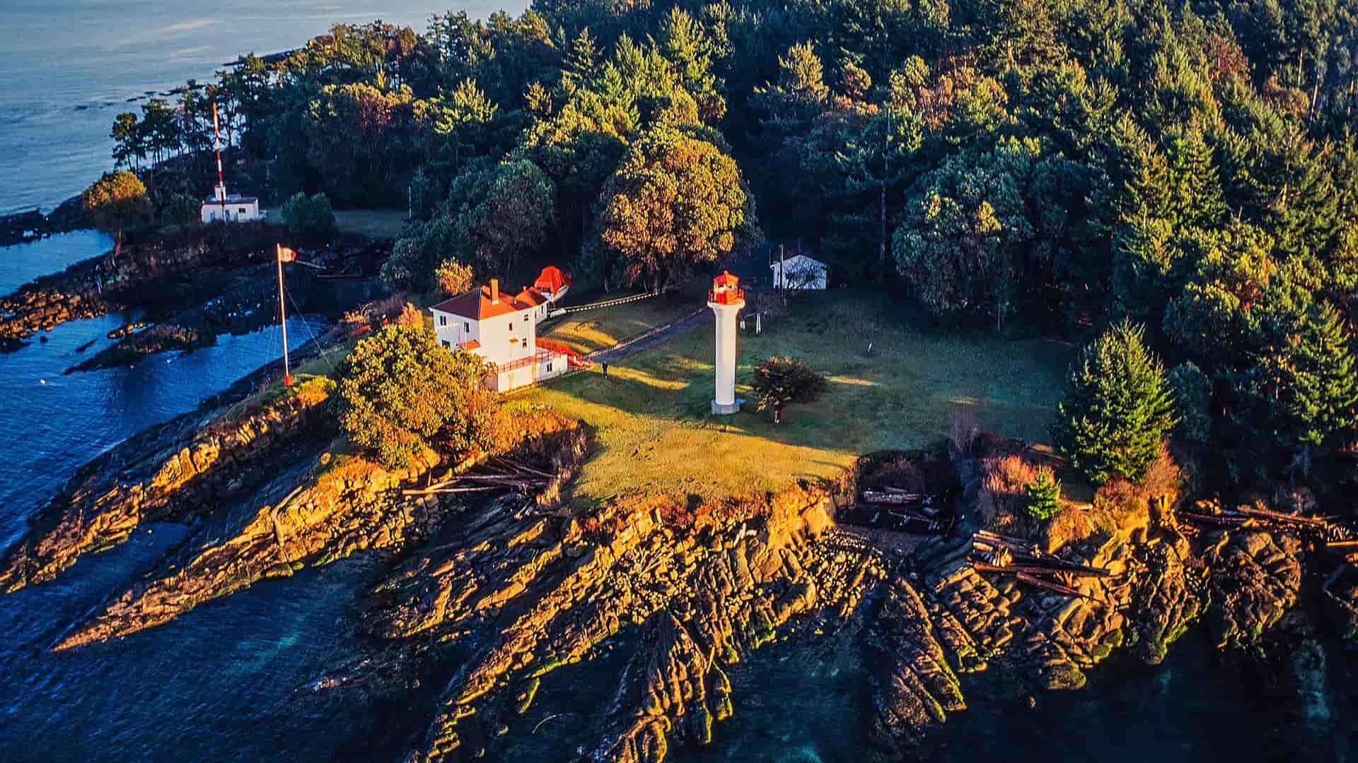 An aerial view of a white lighthouse and residence on a rocky, wooded peninsula jutting into the blue water of Swartz Bay.