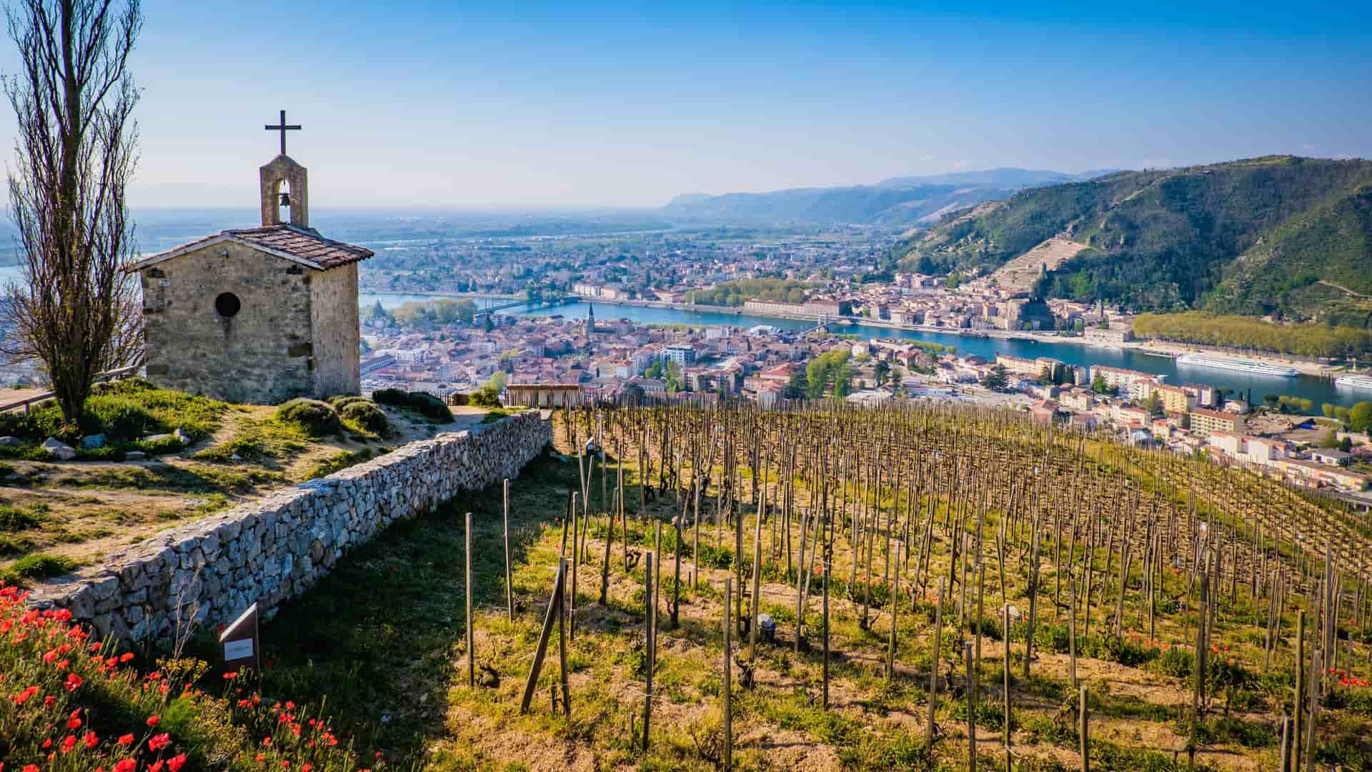 A high-angle view of a vineyard and a small stone chapel with a bell tower in Tain-l'Hermitage, France, overlooking the city and the Rhône River with a bridge spanning it and green mountains in the distance.