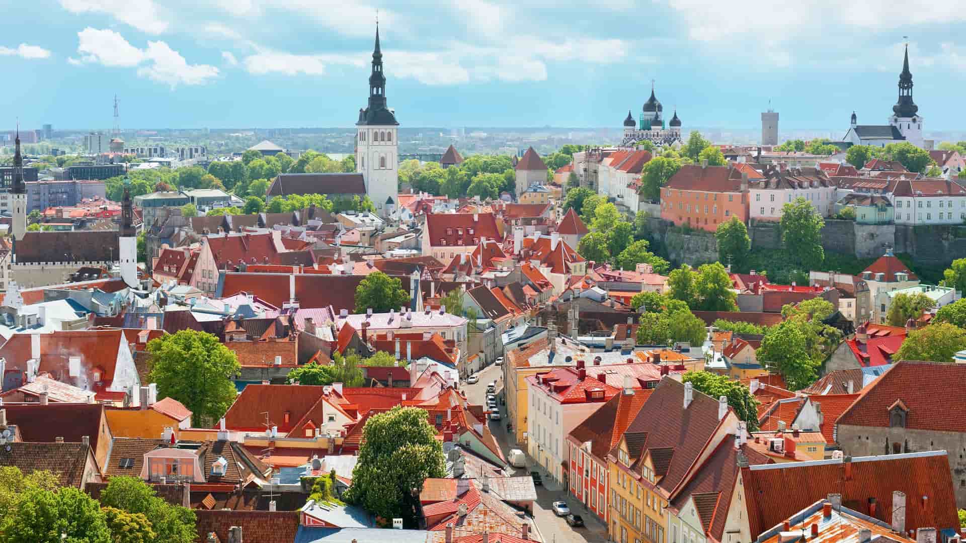 An expansive panoramic view of the medieval Old Town of Tallinn, Estonia, with its iconic red rooftops and historic church spires against a bright, cloudy sky.
