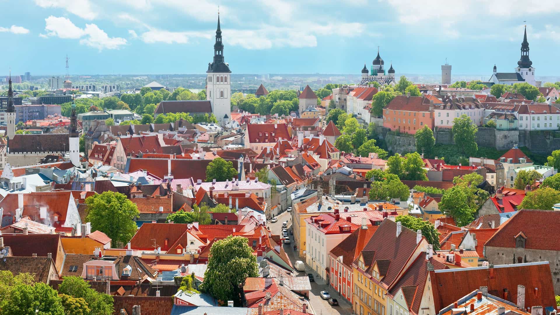 An expansive panoramic view of the medieval Old Town of Tallinn, Estonia, with its iconic red rooftops and historic church spires against a bright, cloudy sky.