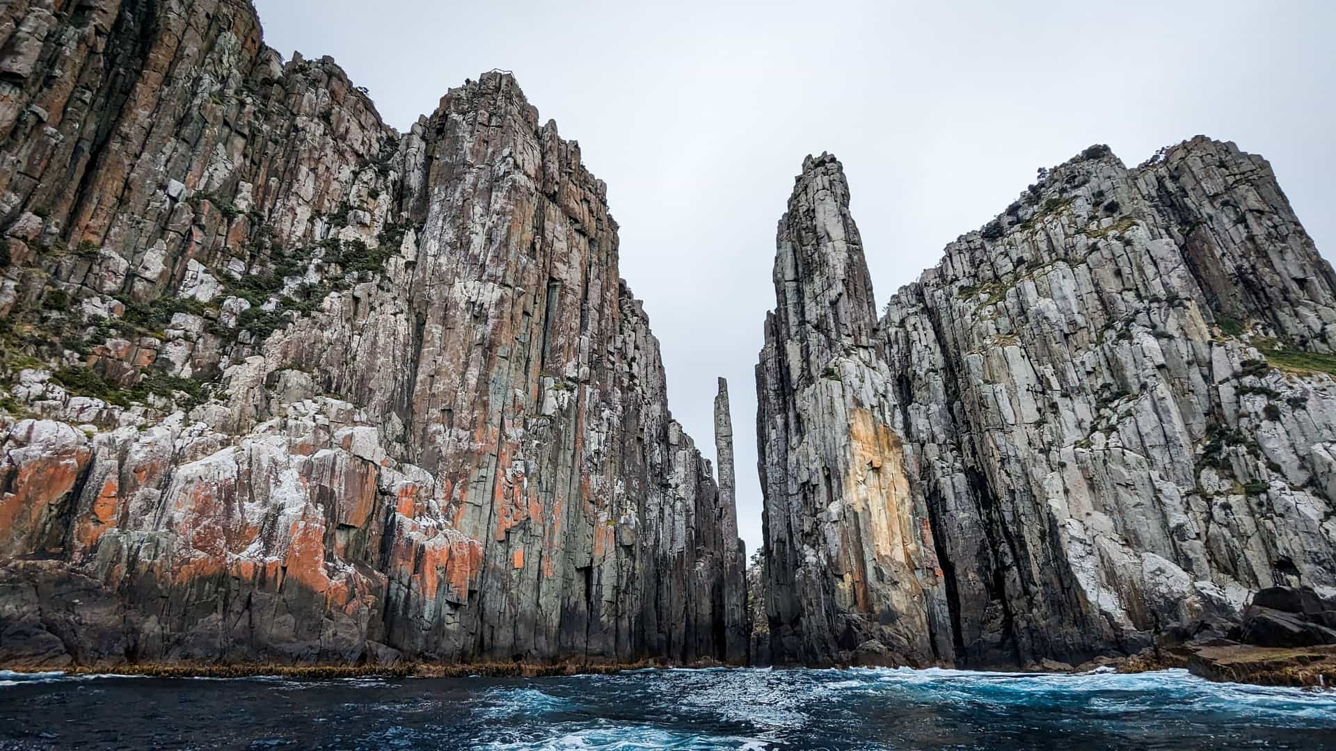 A stunning view of towering, dramatic basalt columns and sea stacks rising from the choppy waters of the Tasman Sea, off the coast of Tasmania, Australia. The sky is overcast and white.