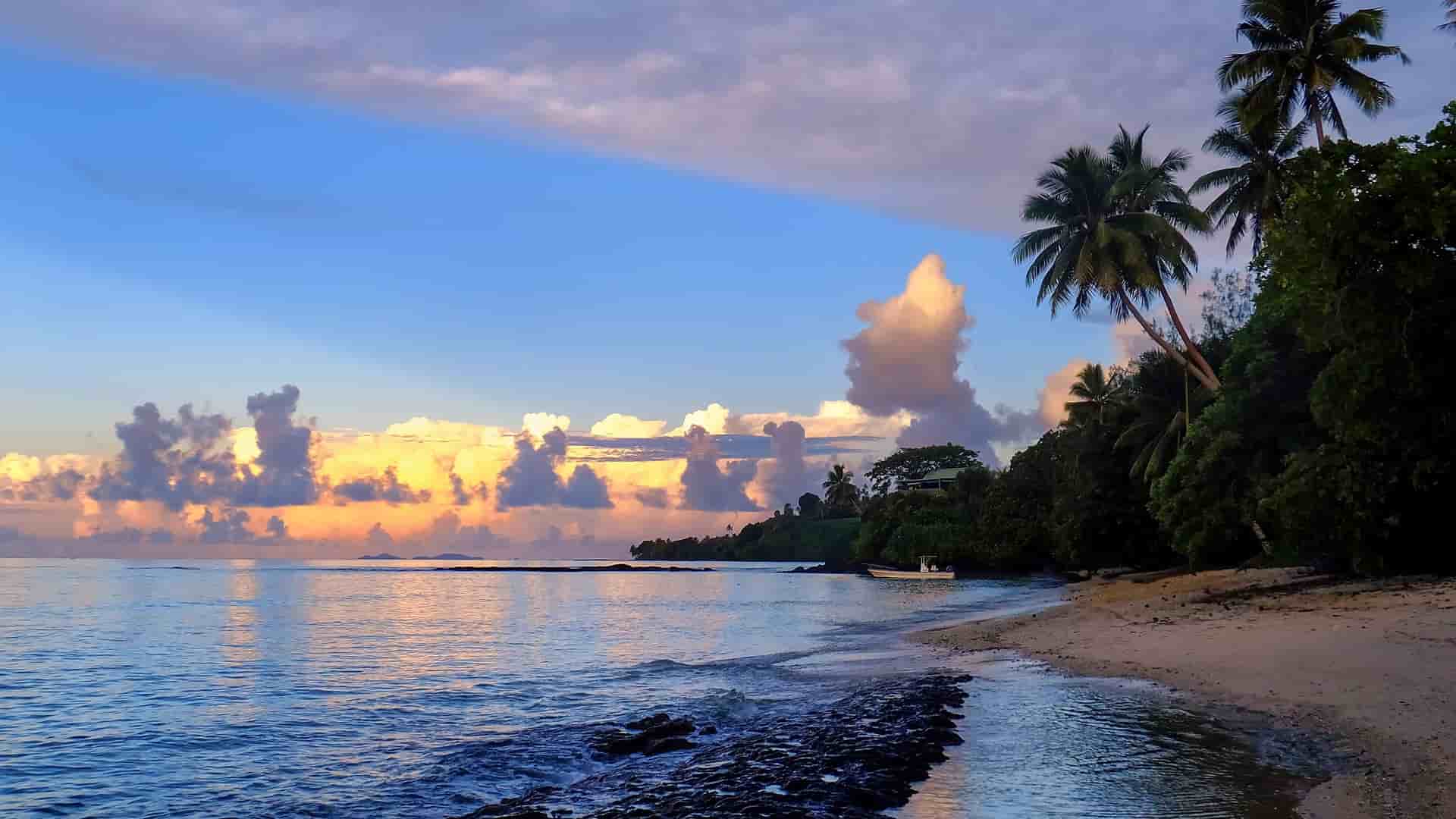 A panoramic landscape of Taveuni Island in Fiji, with a lush, green tropical jungle and palm trees sloping down to a beautiful blue coastline and distant islands under a bright, cloudy sky.