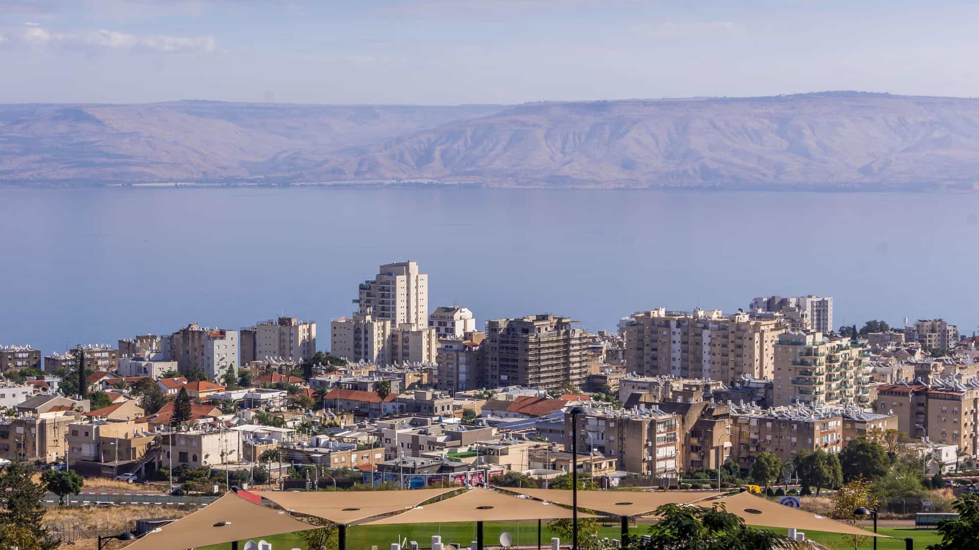 "A scenic view of the city of Tiberias, Israel, showcasing a cluster of buildings along the shore of the calm Sea of Galilee, with rolling desert mountains in the distance under a blue sky. "