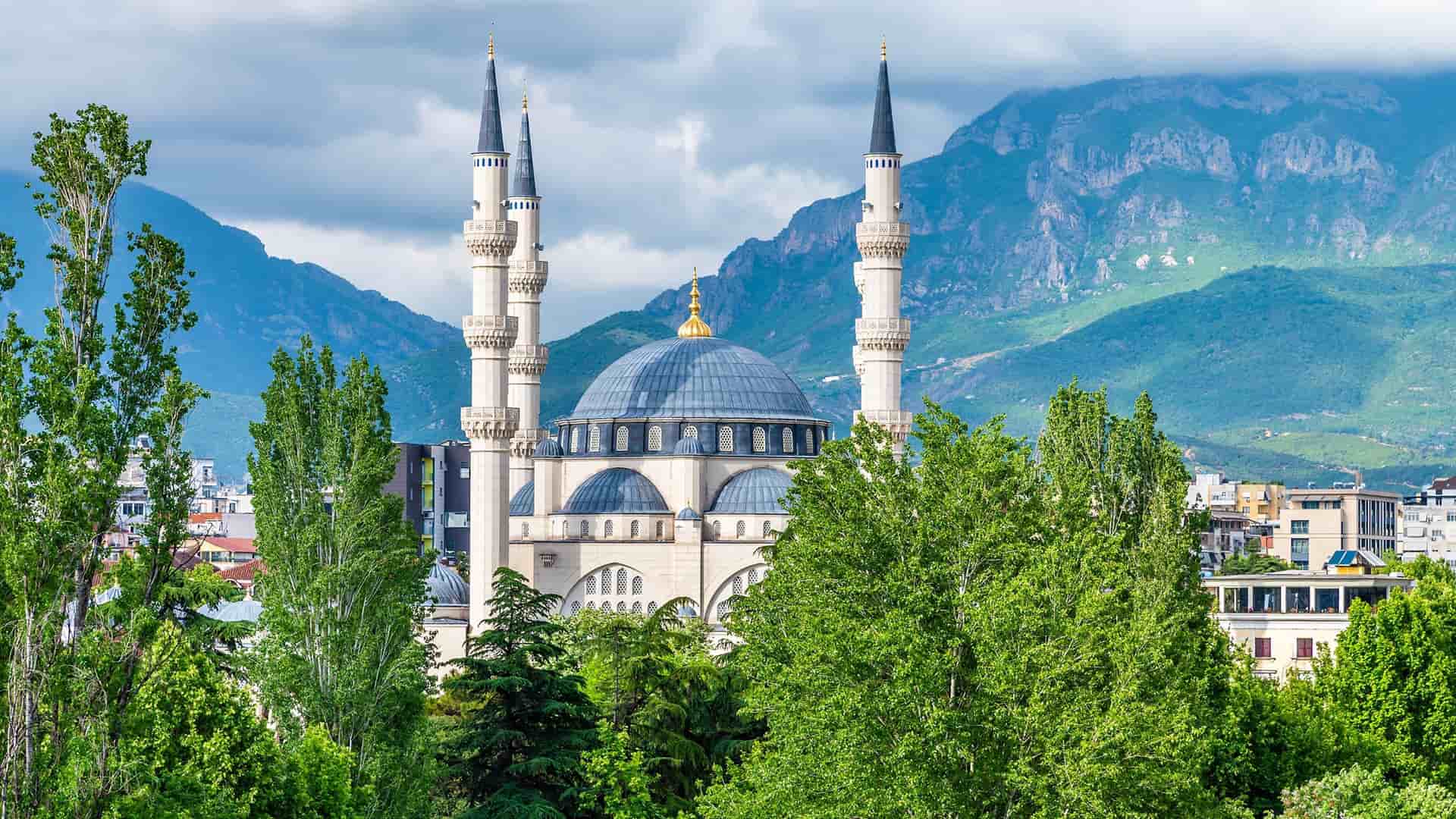 A close-up view of the beautiful blue-domed Namazgjah Mosque in Tirana, Albania, framed by lush green trees with mountains in the background.