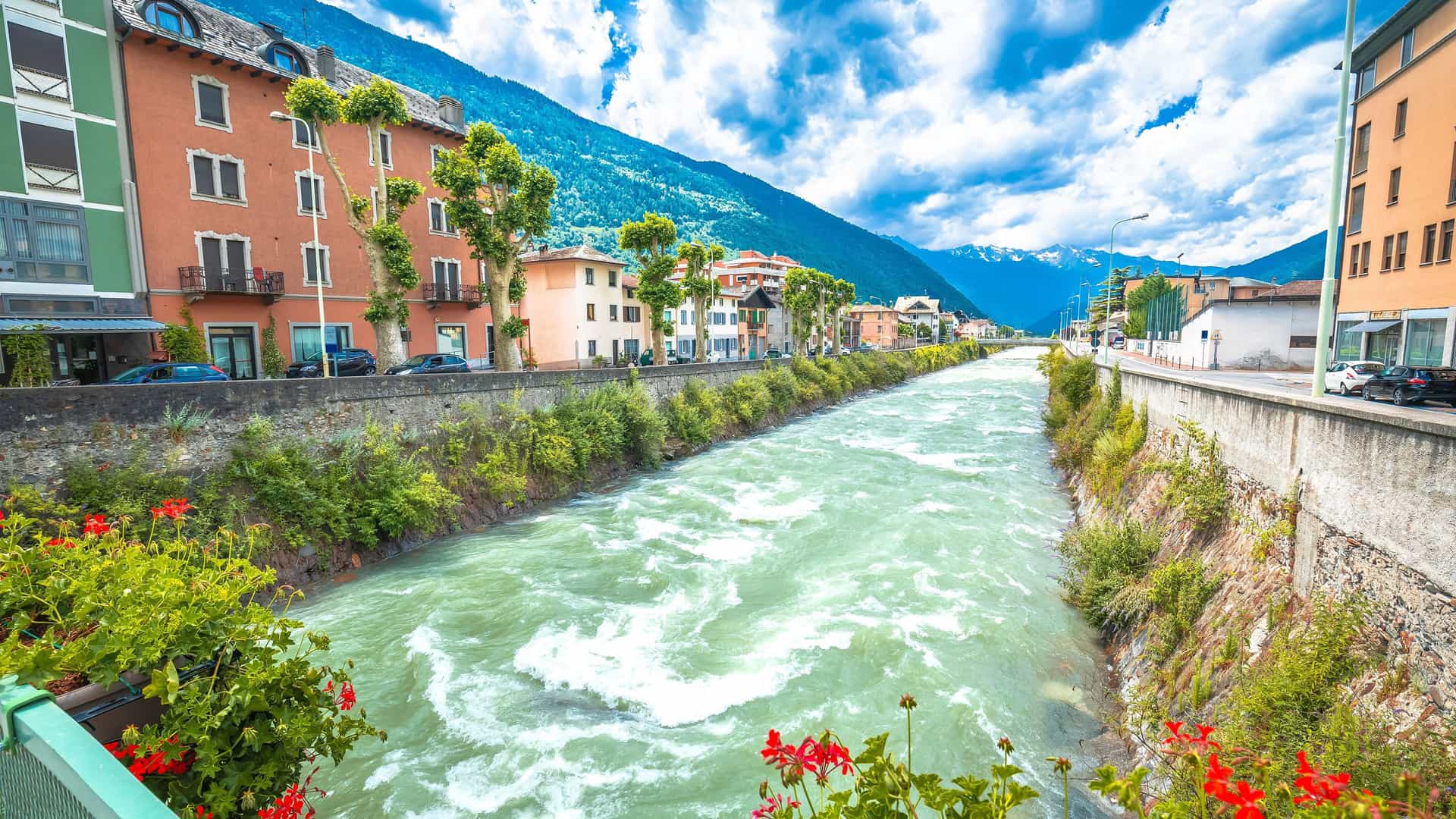 "A picturesque view of the Adda River flowing through the town of Tirano, Italy, with colorful historic buildings and the dramatic Italian Alps in the background. "