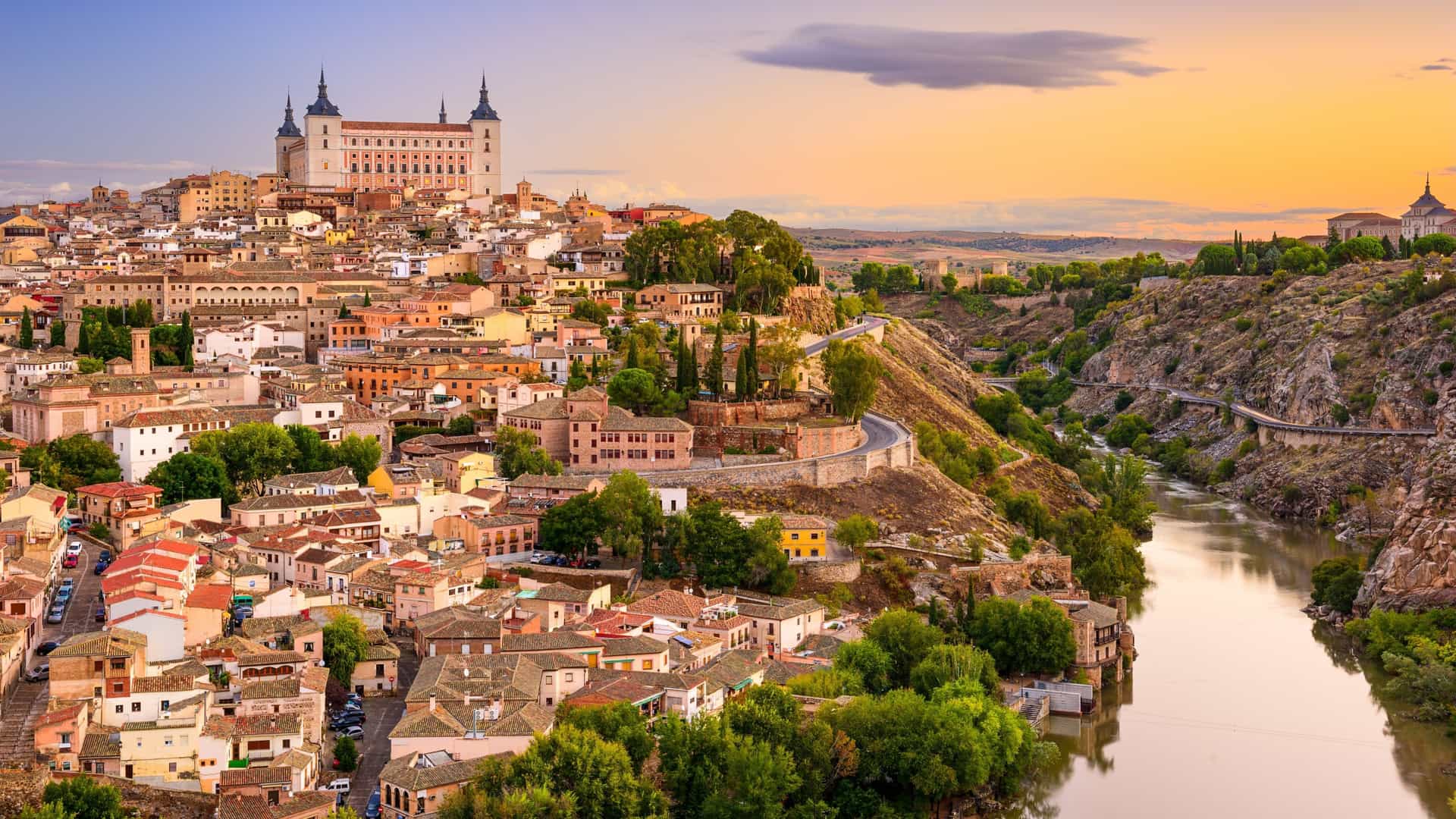 A panoramic view of the UNESCO World Heritage city of Toledo, Spain. The historic, old city is dramatically perched on a hill, with the massive Alcázar fortress and the spire of the Toledo Cathedral dominating the skyline. In the foreground, the Tagus River (Río Tajo) winds around the base of the rock where the city is built.