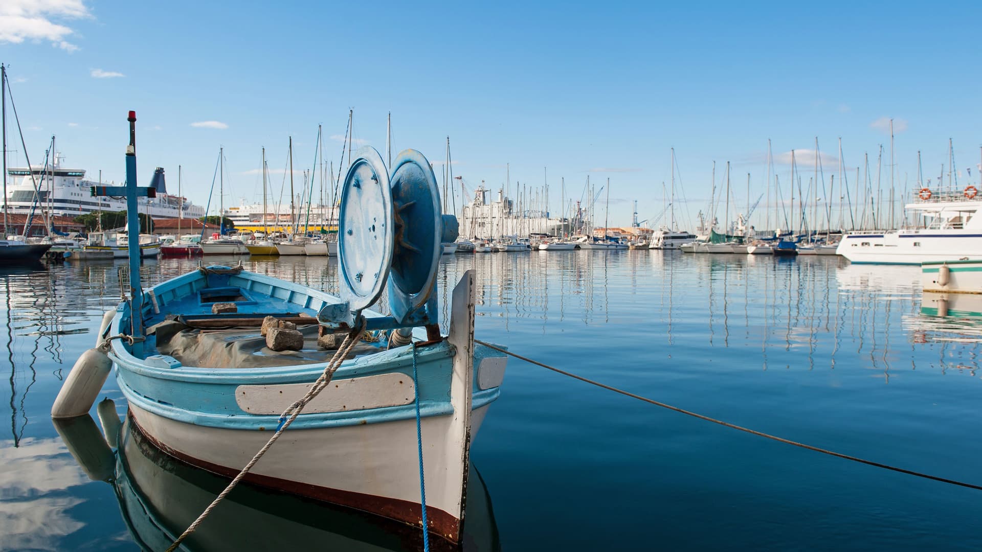 A classic blue fishing boat docked in the busy port of Toulon, France, with numerous sailboats and yachts reflected in the calm water.