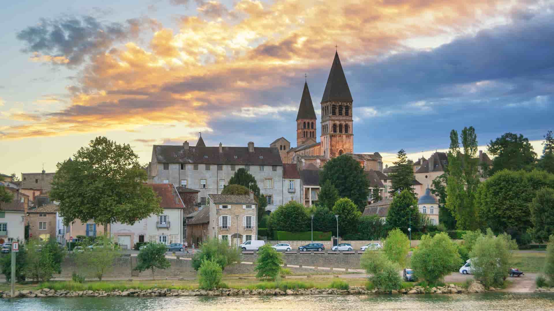 A panoramic view of the historic town of Tournus, France, with its famous Abbey of Saint Philibert and other buildings along the SaƓne River at sunset.
