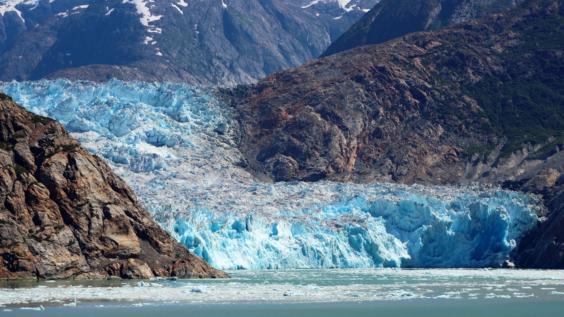 A panoramic view of a massive blue and white glacier in the Tracy Arm Fjord, with jagged rocky cliffs and a field of floating ice chunks in the water.