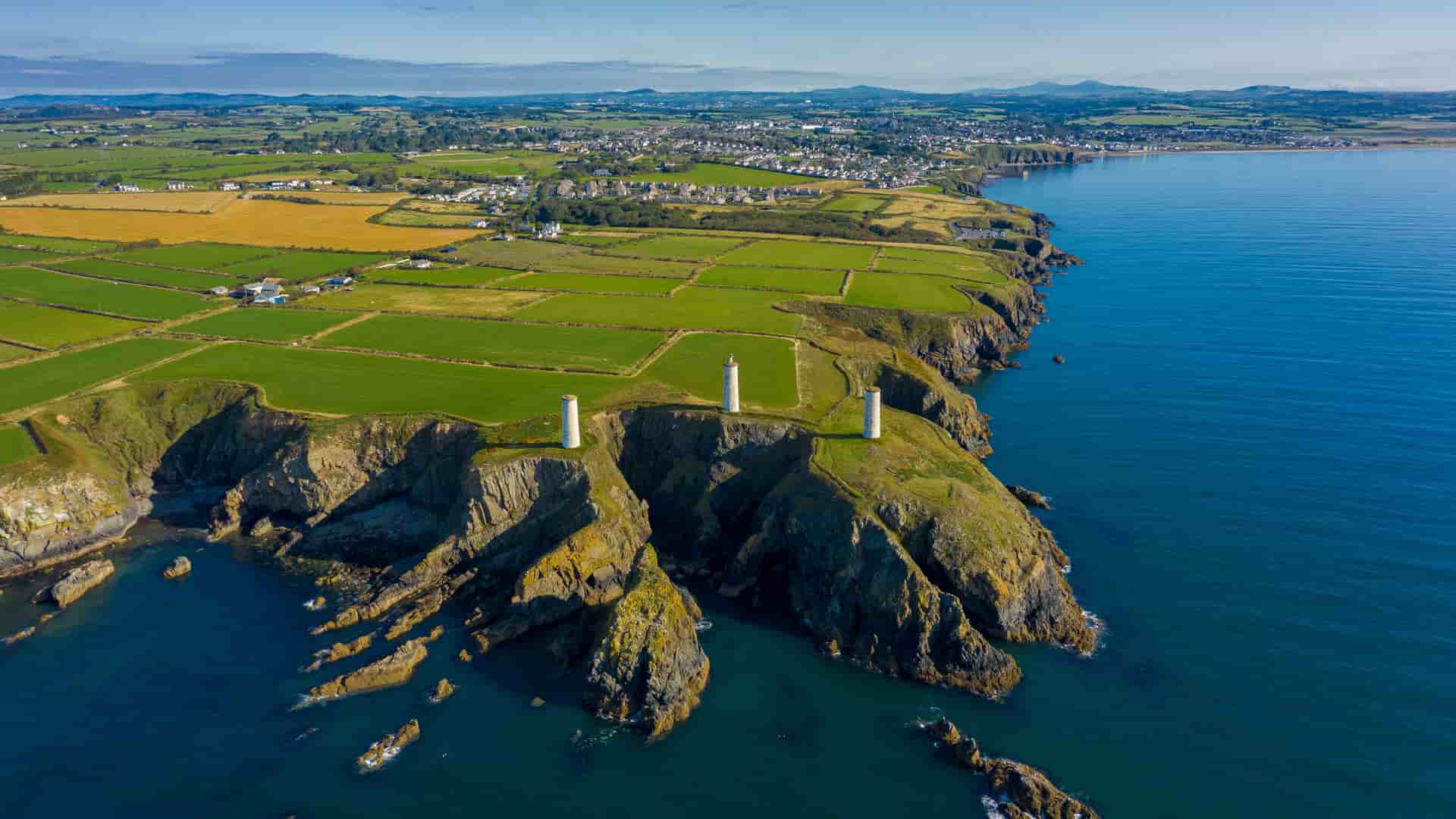 "An aerial view of the rugged coastline of Tramore, Ireland, showcasing the three historic stone pillars standing on the cliff's edge, overlooking the deep blue sea. The surrounding landscape features a patchwork of green and yellow fields leading to a small town in the distance. "