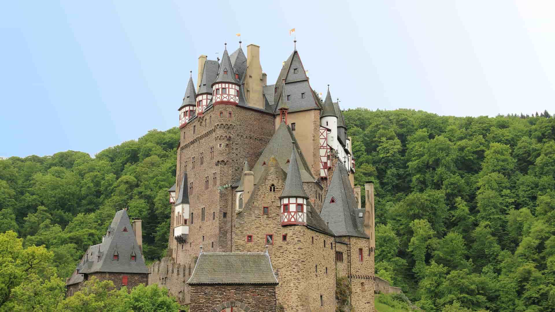 A majestic Eltz Castle, a medieval landmark with multiple towers and red-framed timber sections, nestled in a lush, dense forest near Trier, Germany.