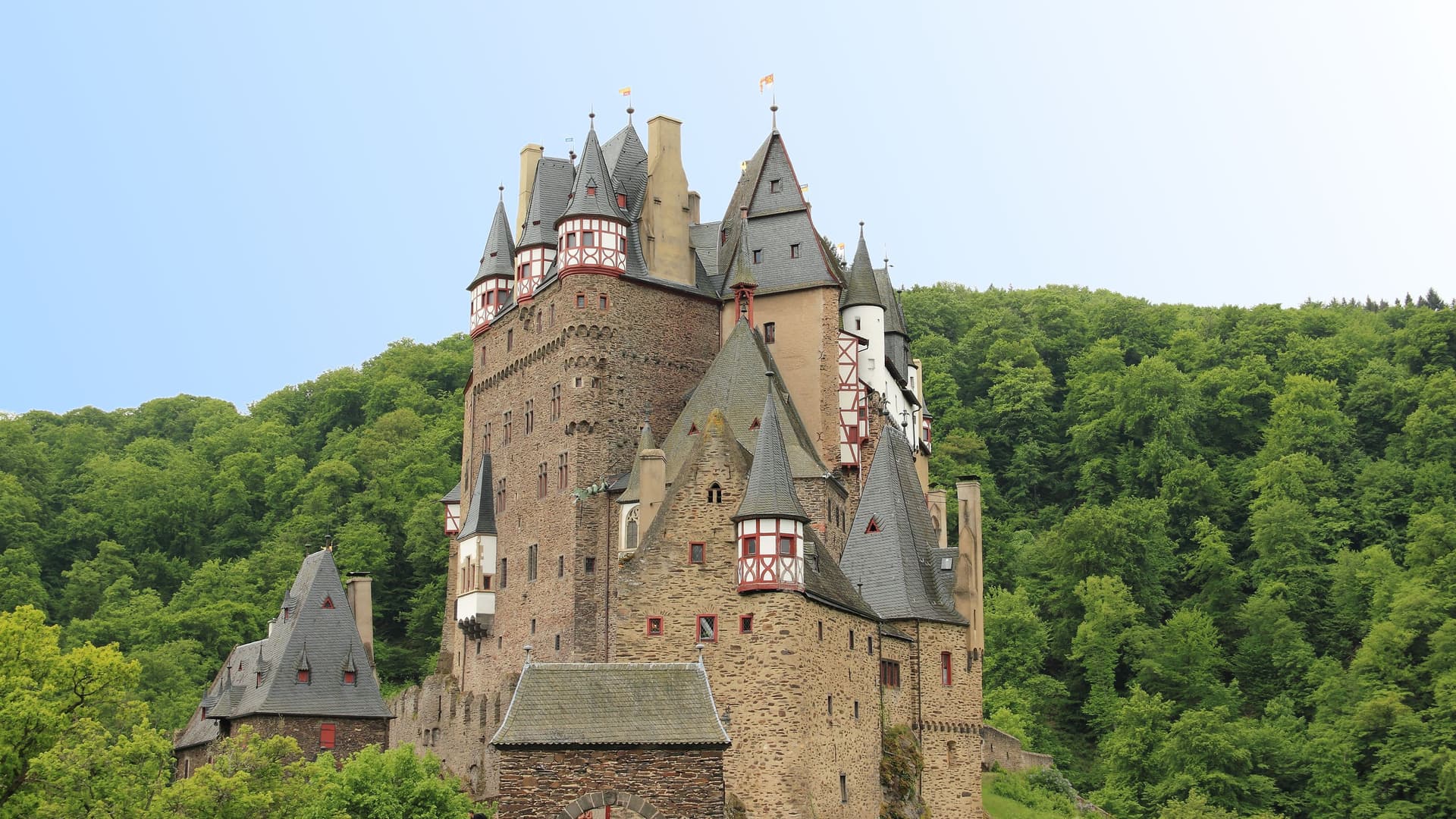 A majestic Eltz Castle, a medieval landmark with multiple towers and red-framed timber sections, nestled in a lush, dense forest near Trier, Germany.