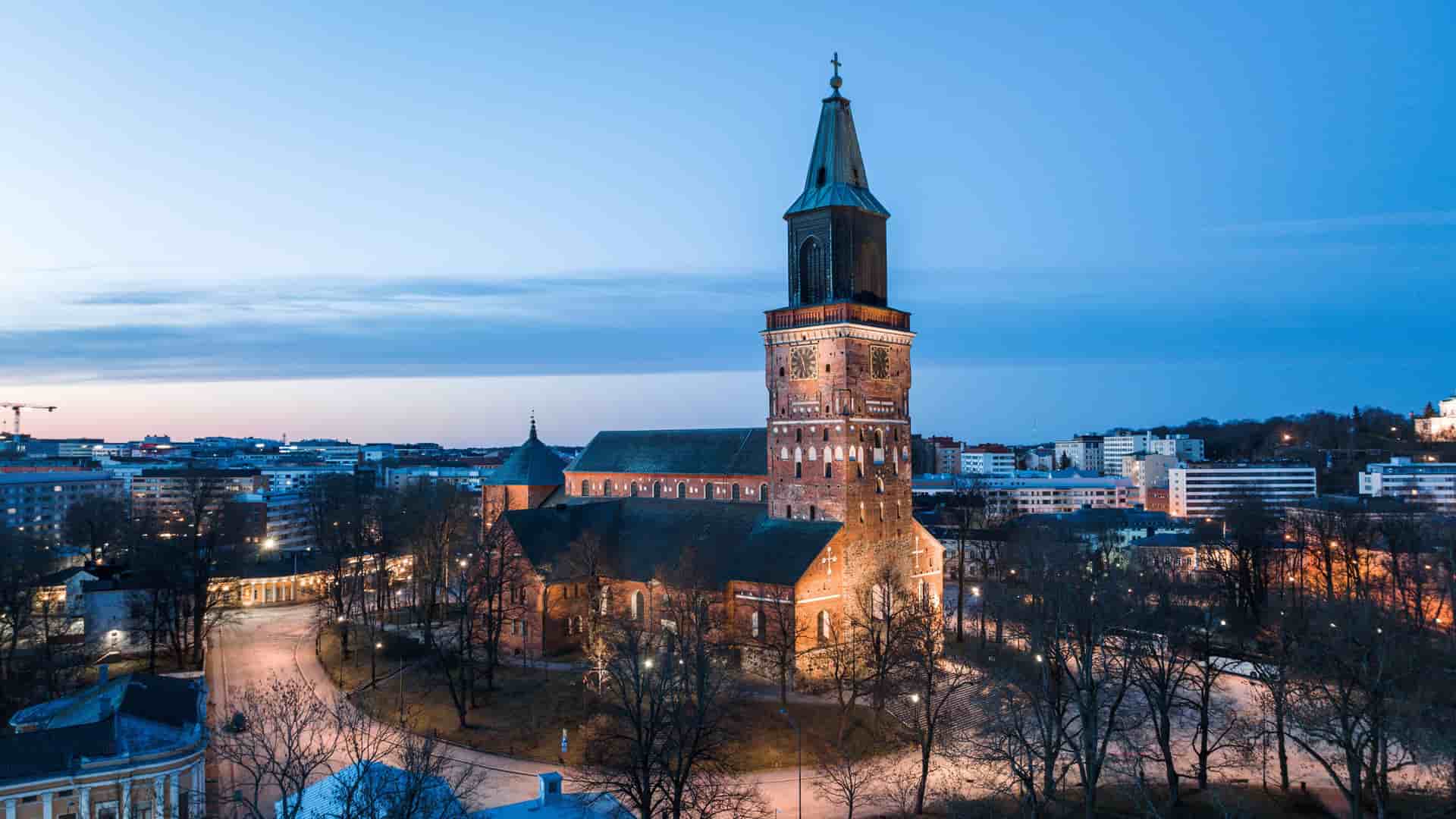 An aerial shot of the medieval Turku Cathedral illuminated at dusk, its brick exterior and tall spire a prominent landmark in the Finnish cityscape.