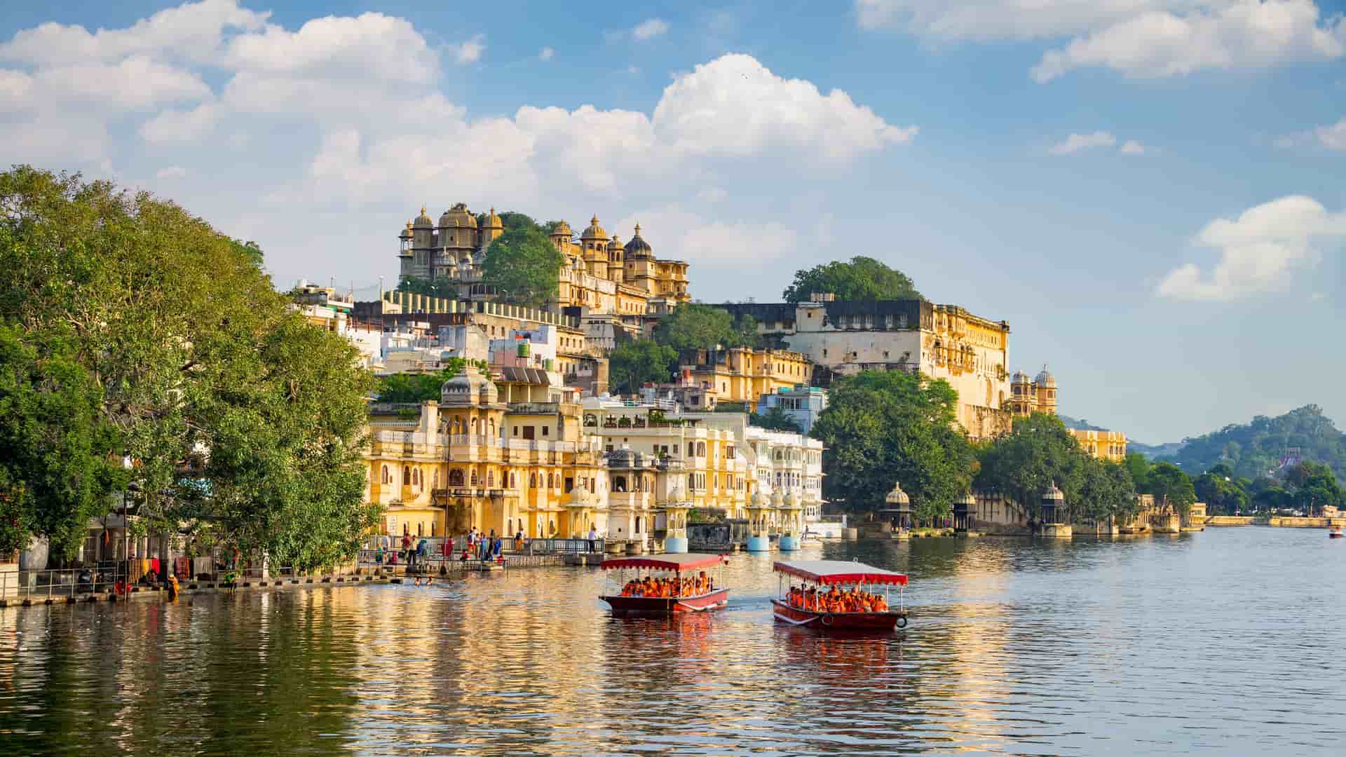 A picturesque view of the historic city of Udaipur, India, with the City Palace and other traditional buildings on the shores of Lake Pichola with two boats sailing on it.