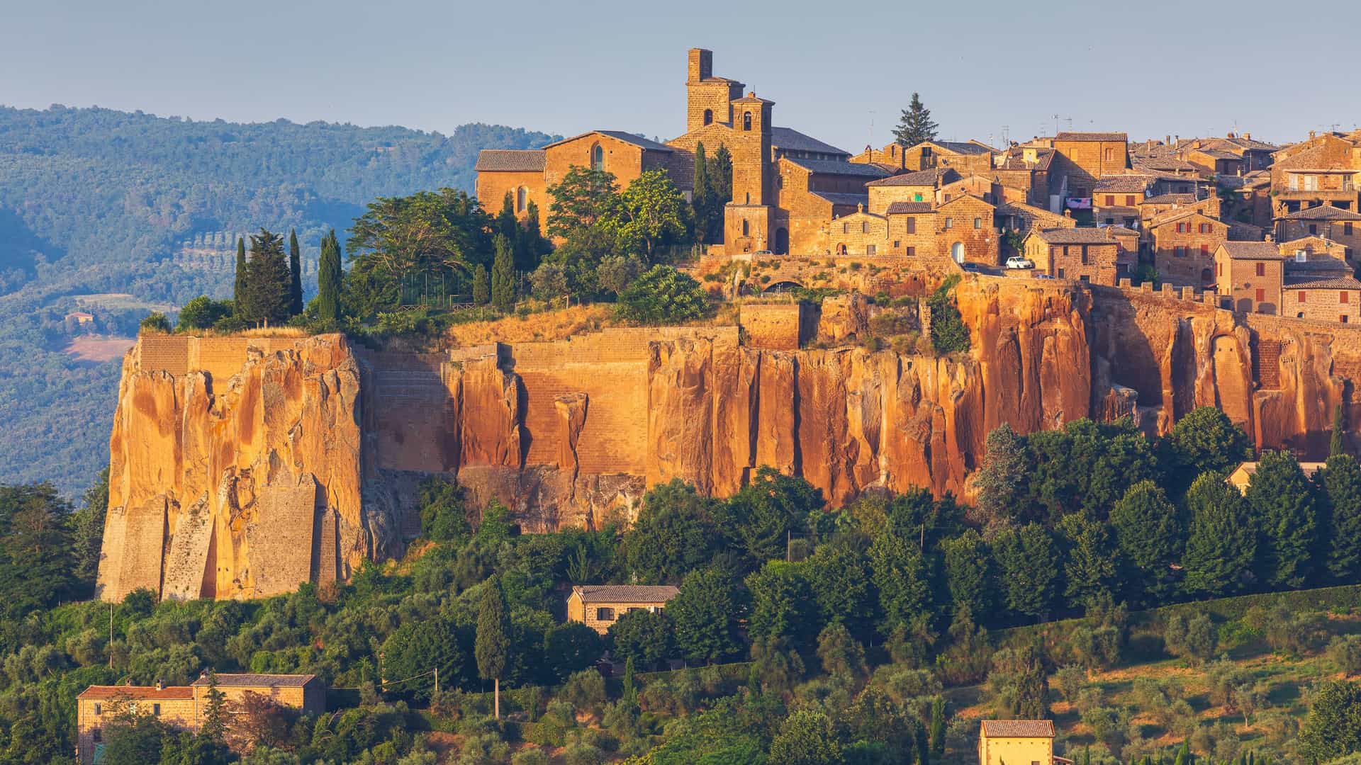 "A beautiful long-distance shot of the medieval town of Orvieto in Umbria, Italy, perched dramatically atop a high volcanic rock cliff, glowing in the late afternoon sun. "