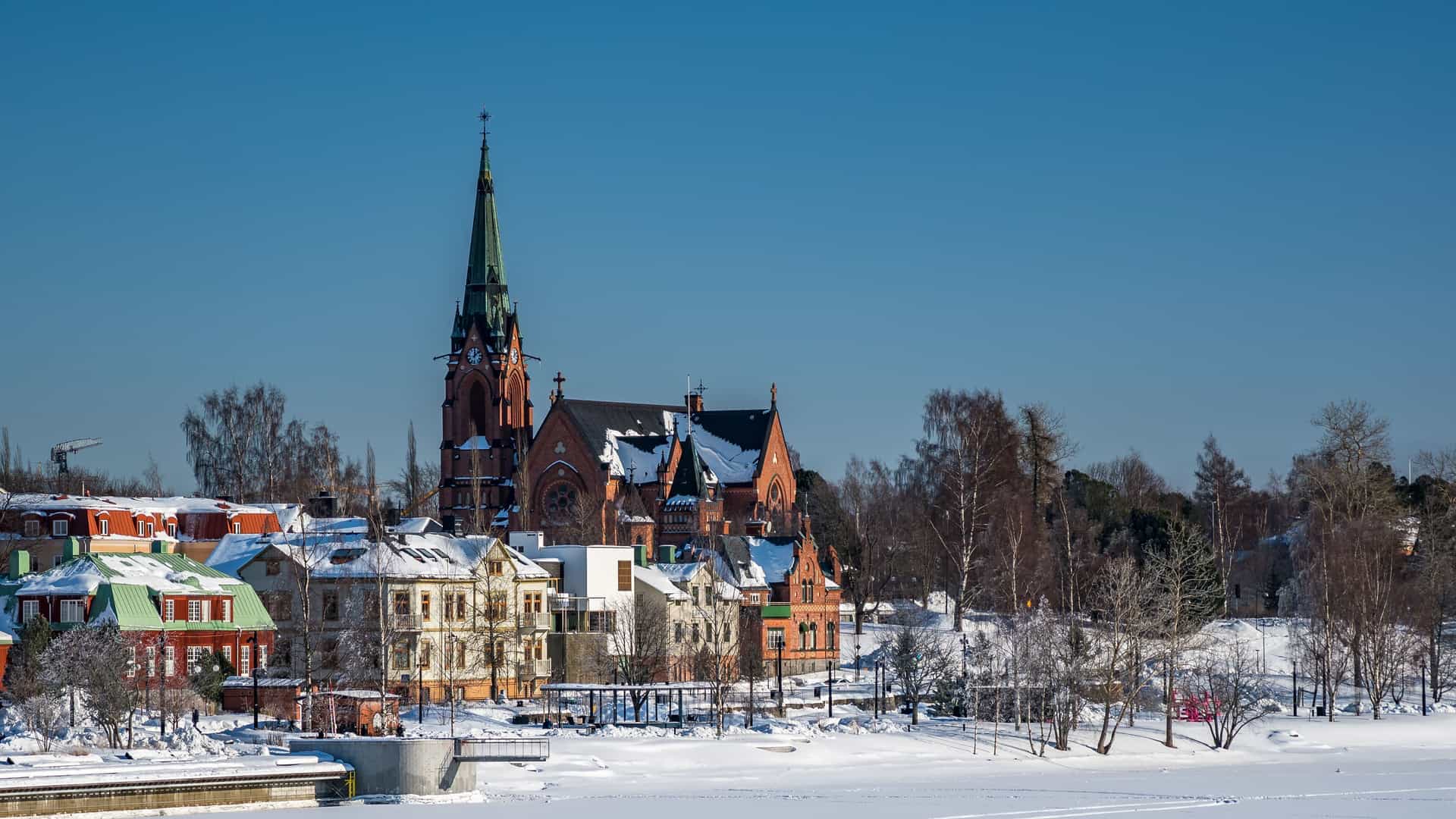 A scenic winter view of the red brick Umeå City Church, its towering spire rising above snow-covered buildings along the frozen Ume River in Sweden.