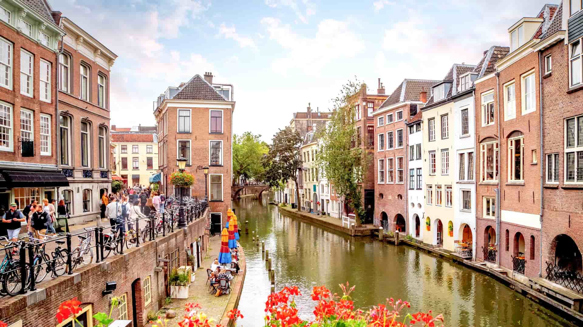 A beautiful canal view in Utrecht, Netherlands, with historic houses, a busy wharf with cafes, and colorful flowers, all under a bright blue sky.