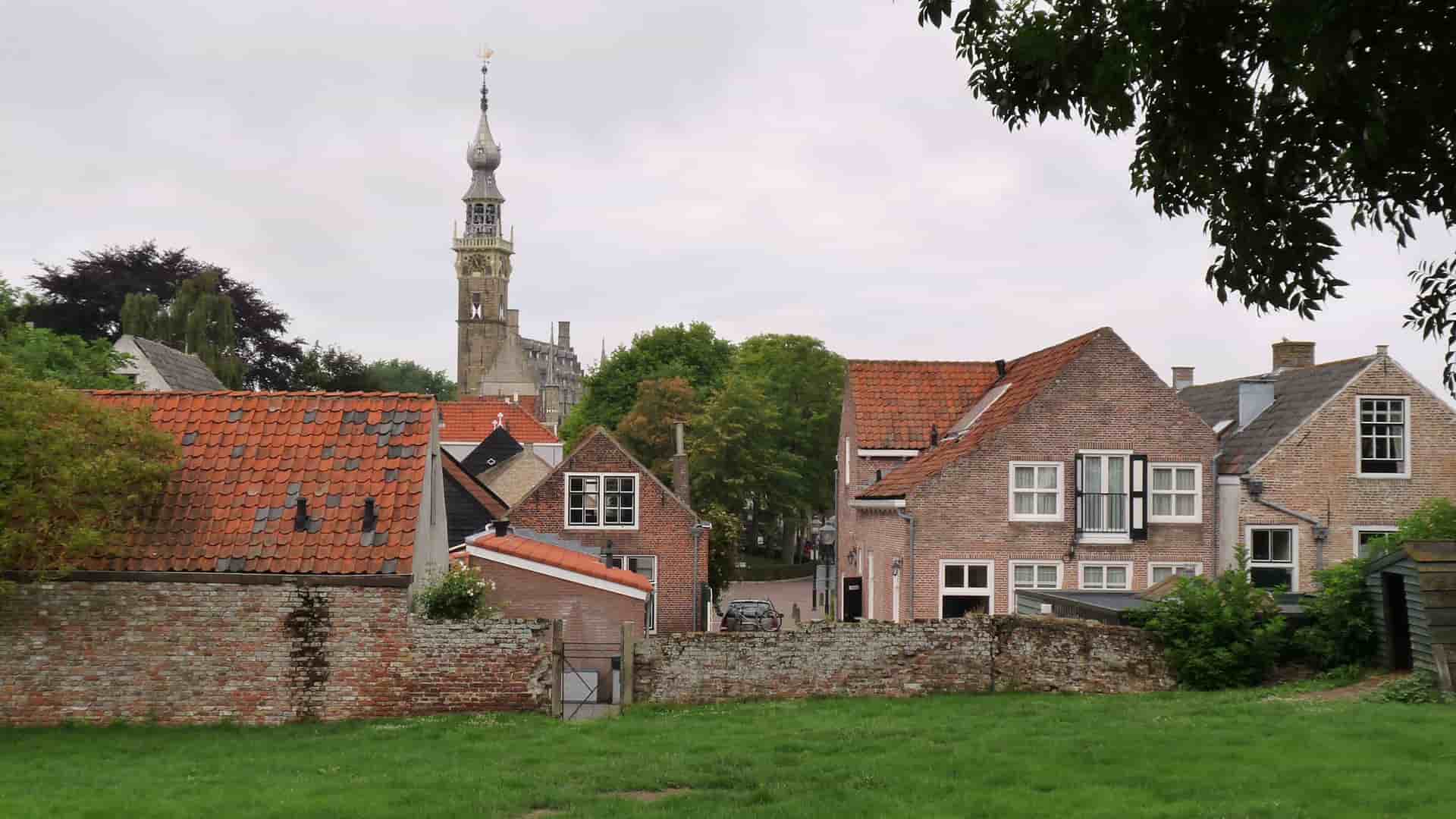 A row of historic red brick houses with red tiled roofs, a stone wall, and a church steeple in the background in the quaint town of Veere, Netherlands.