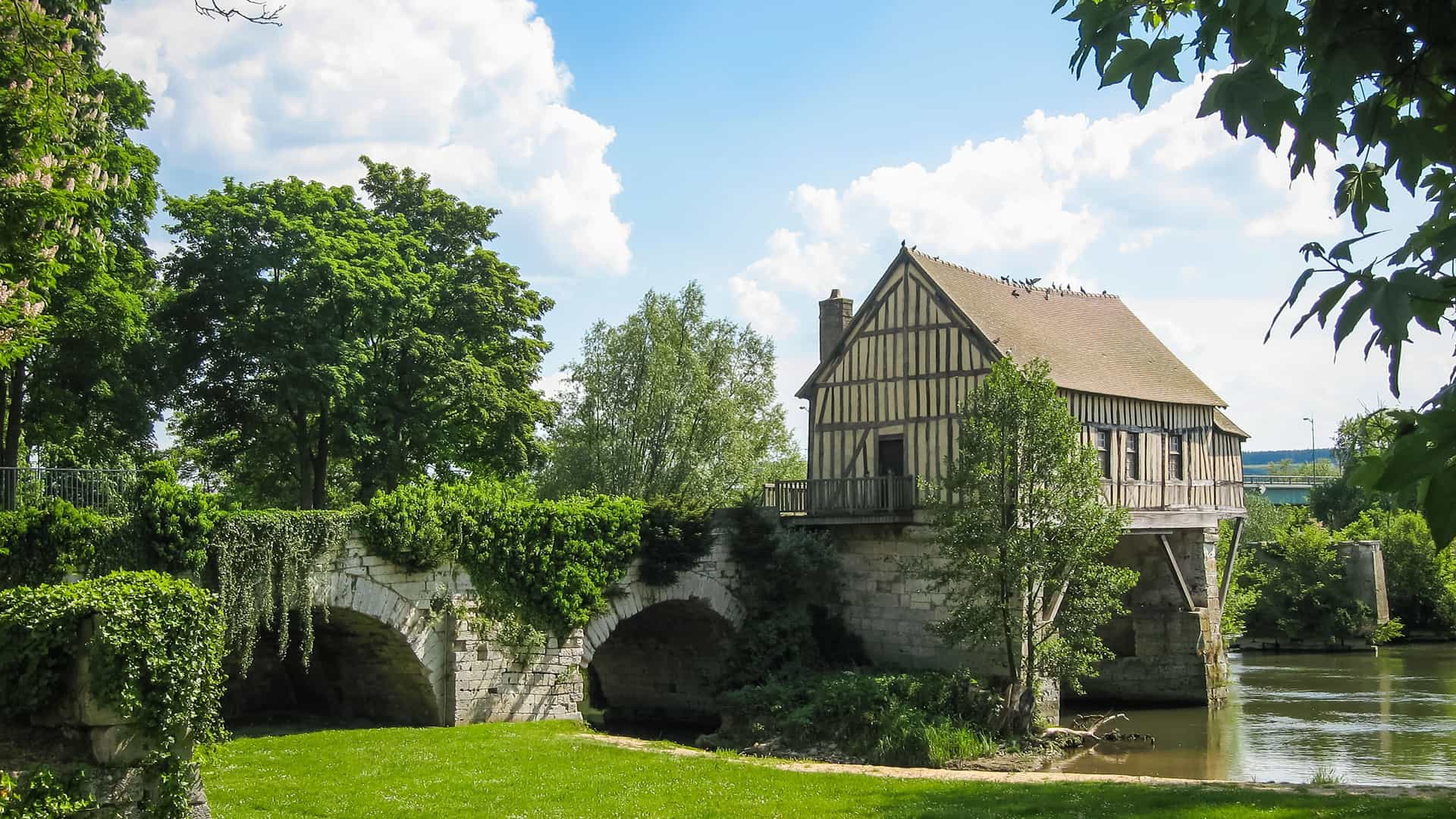 A classic half-timbered house with a light roof, situated on a stone bridge over a calm river in Vernon, France, surrounded by lush green trees and foliage.