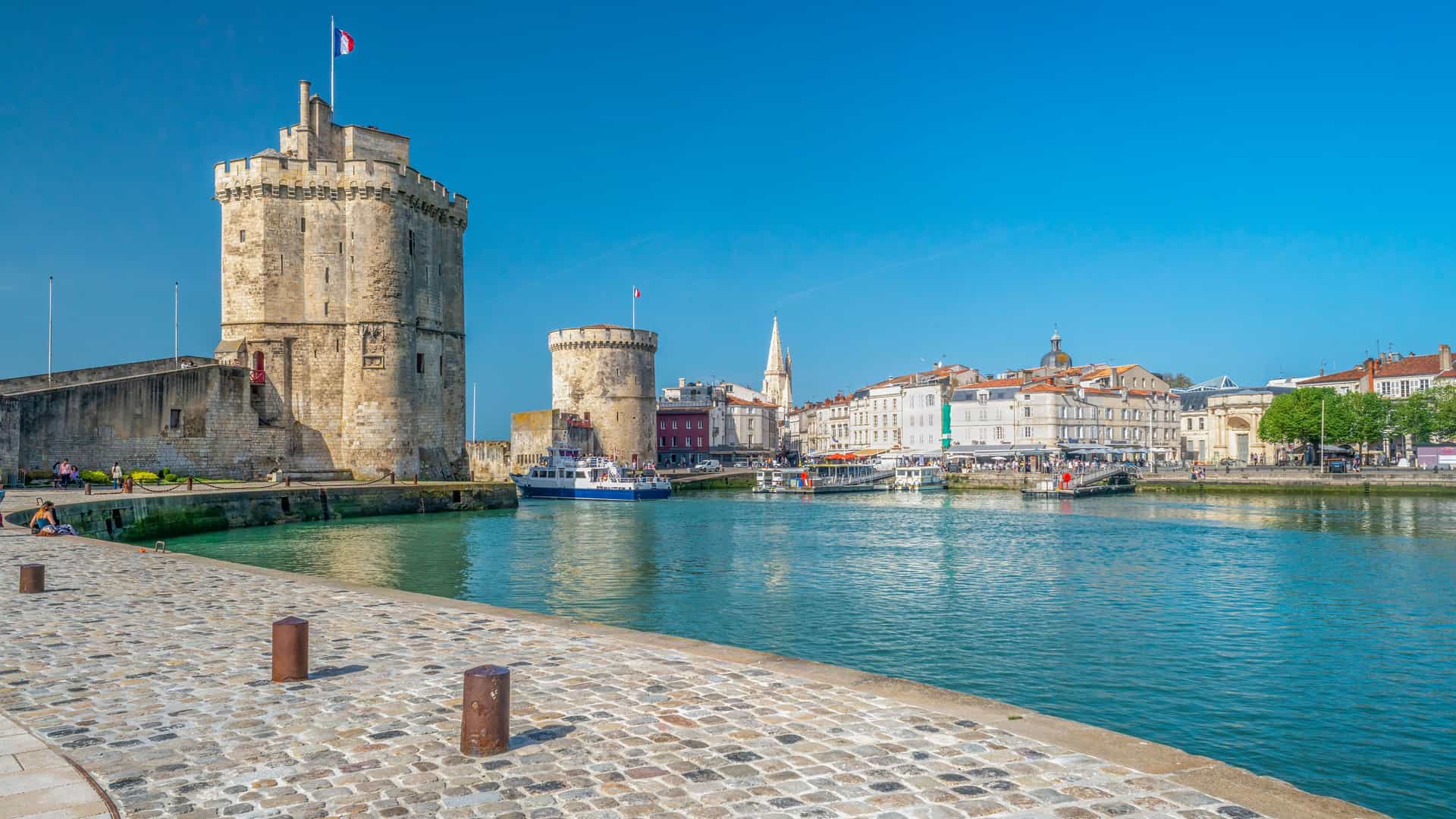 A vibrant harborside view of the historic medieval Towers of La Rochelle, a major landmark, with a cobblestone quay and boats floating in the calm water on a sunny day.
