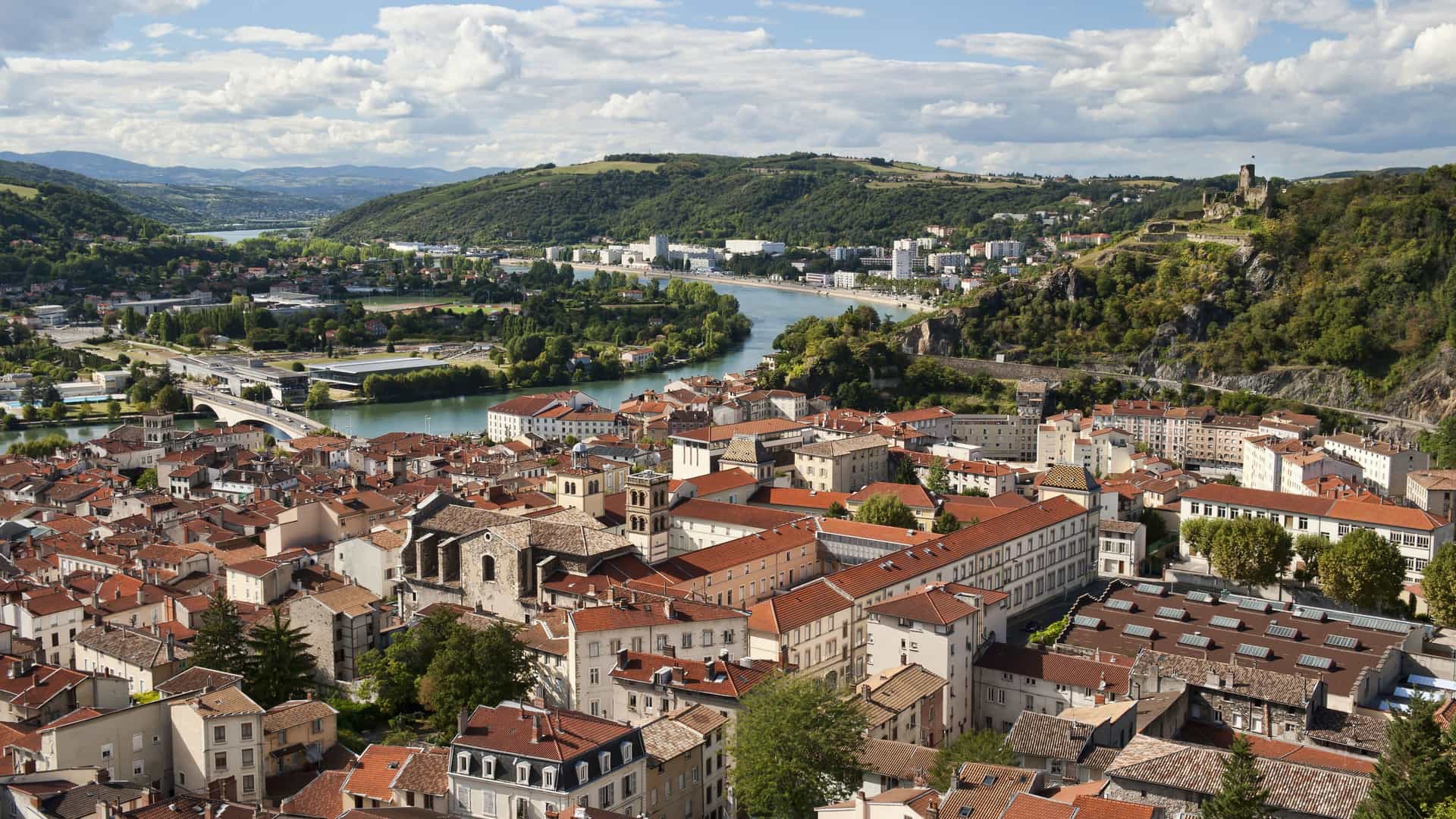 An expansive aerial view of the historic city of Vienne, France, with its red-tiled roofs and the Rhone River winding through the valley surrounded by green, rolling hills and a castle ruin on the hilltop.