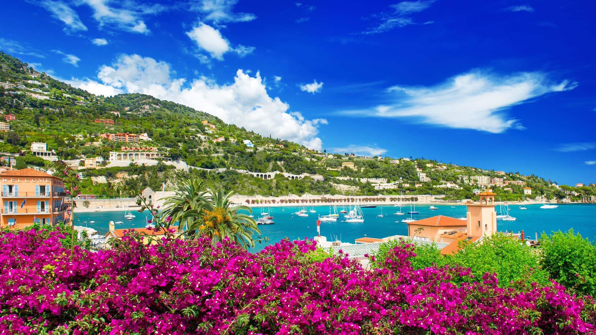 A vibrant view of the picturesque town of Villefranche-sur-Mer, France, with colorful buildings and yachts in the bay, set against a backdrop of steep, green hills under a bright blue sky.