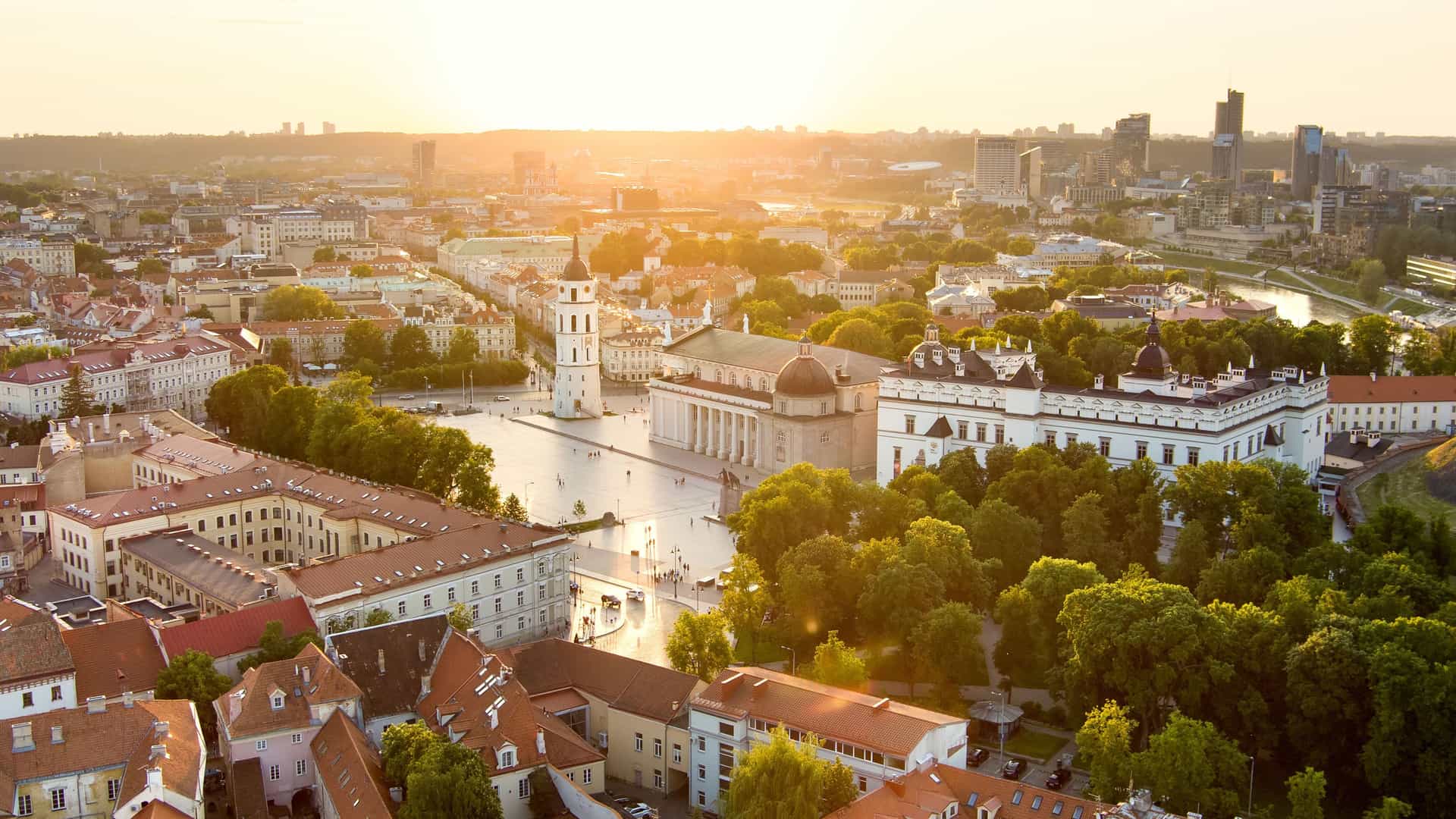 "An aerial view of Vilnius, Lithuania, at sunset, showcasing the historic Vilnius Cathedral and its prominent bell tower in Cathedral Square, with the modern cityscape and the Neris River in the background. "