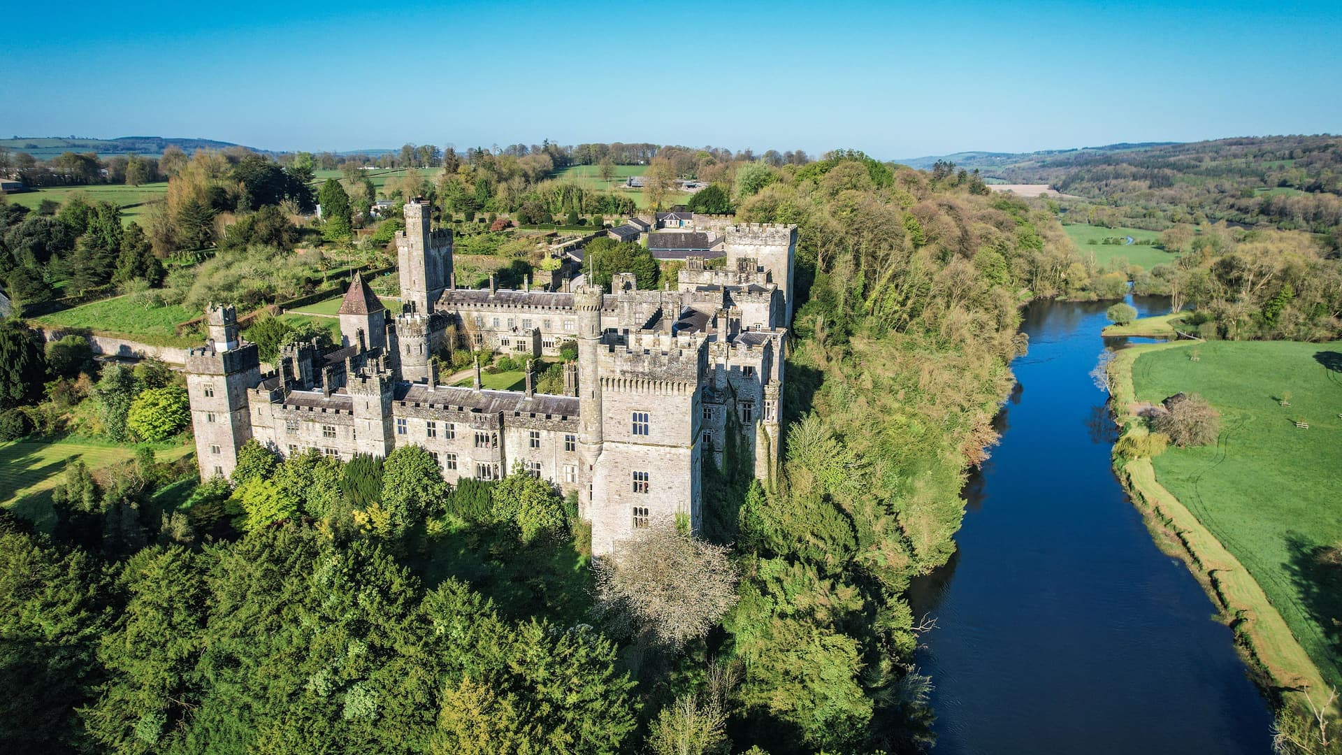 An aerial view of the historic Lismore Castle in County Waterford, Ireland, located on a bank of the River Blackwater and surrounded by lush green forests.