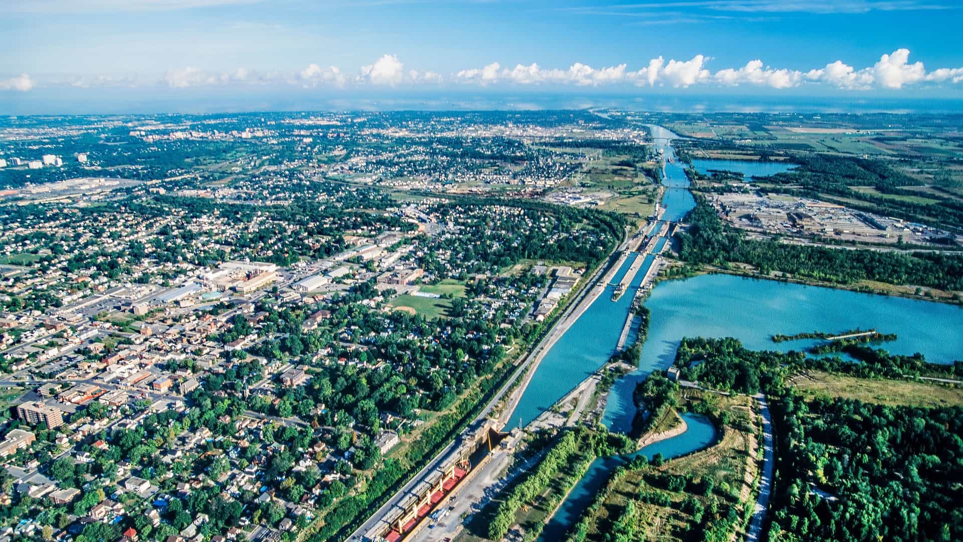 An aerial view of a cargo ship navigating the Welland Canal, with the locks and surrounding parkland, separating the city and Lake Ontario from the sprawling suburbs and industrial areas.