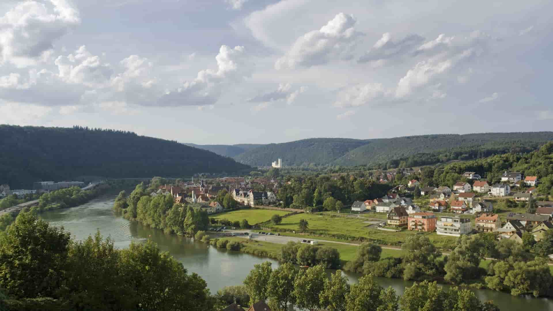 A high-angle view of the charming town of Wertheim, Germany, nestled on the banks of the Main River, surrounded by lush, green hills and a scenic valley.