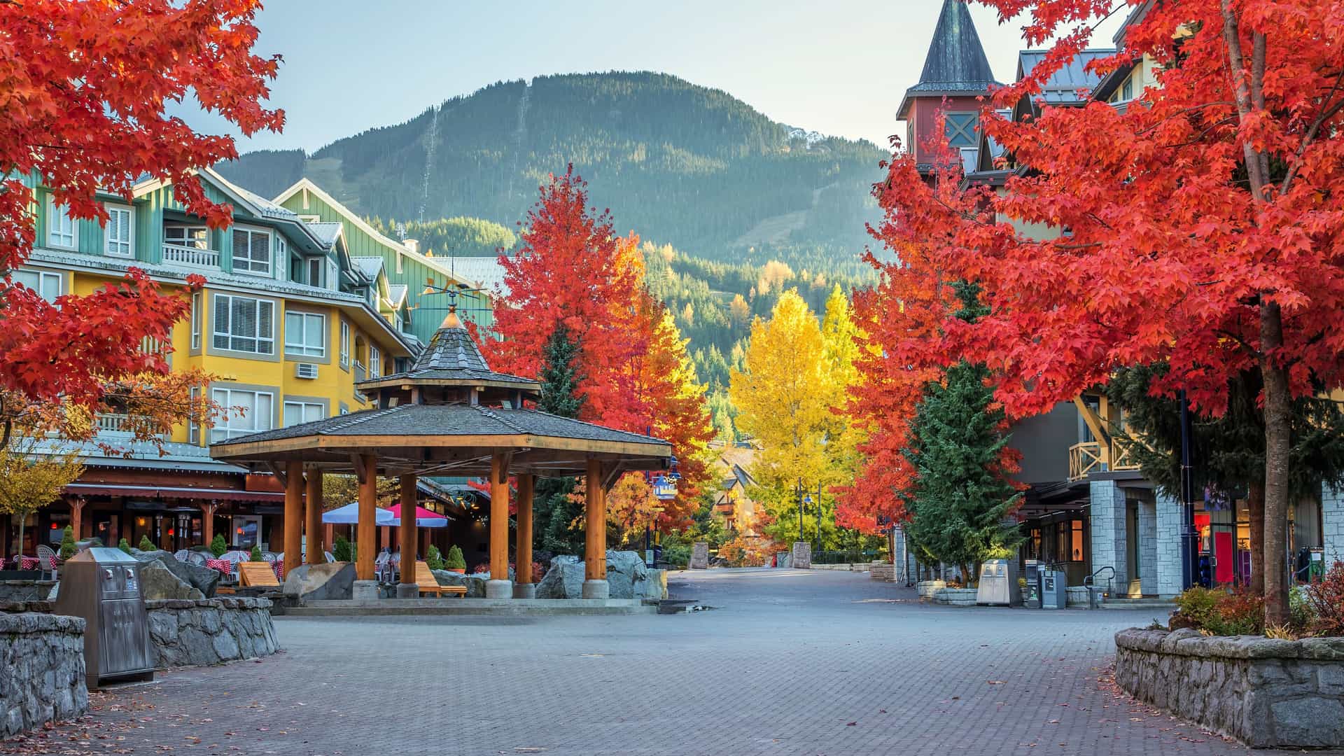"A scenic view of Whistler Village Square in autumn, with colorful buildings, a central gazebo, and trees with vibrant red and yellow foliage against a backdrop of mountains. "