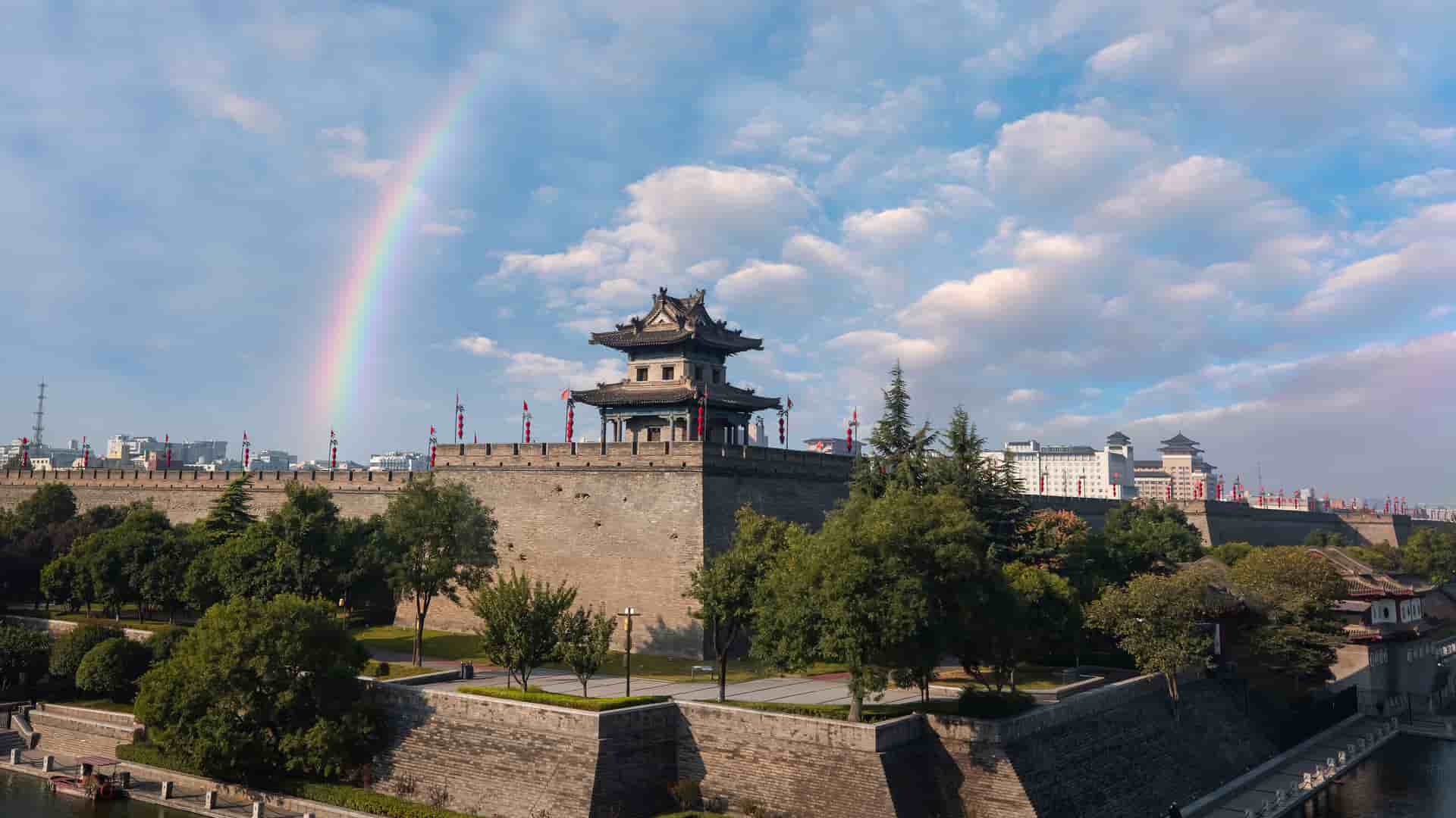 A vibrant rainbow arcs over the historic City Wall in Xi'an, China, a large defensive stone structure with a traditional Chinese watchtower and surrounding moat.