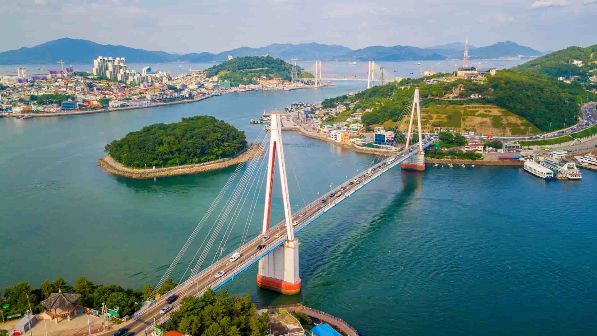 An aerial view of the Dolsan Bridge, a modern cable-stayed bridge in Yeosu, South Korea, connecting Dolsan Island to the mainland with a city and mountains in the background.