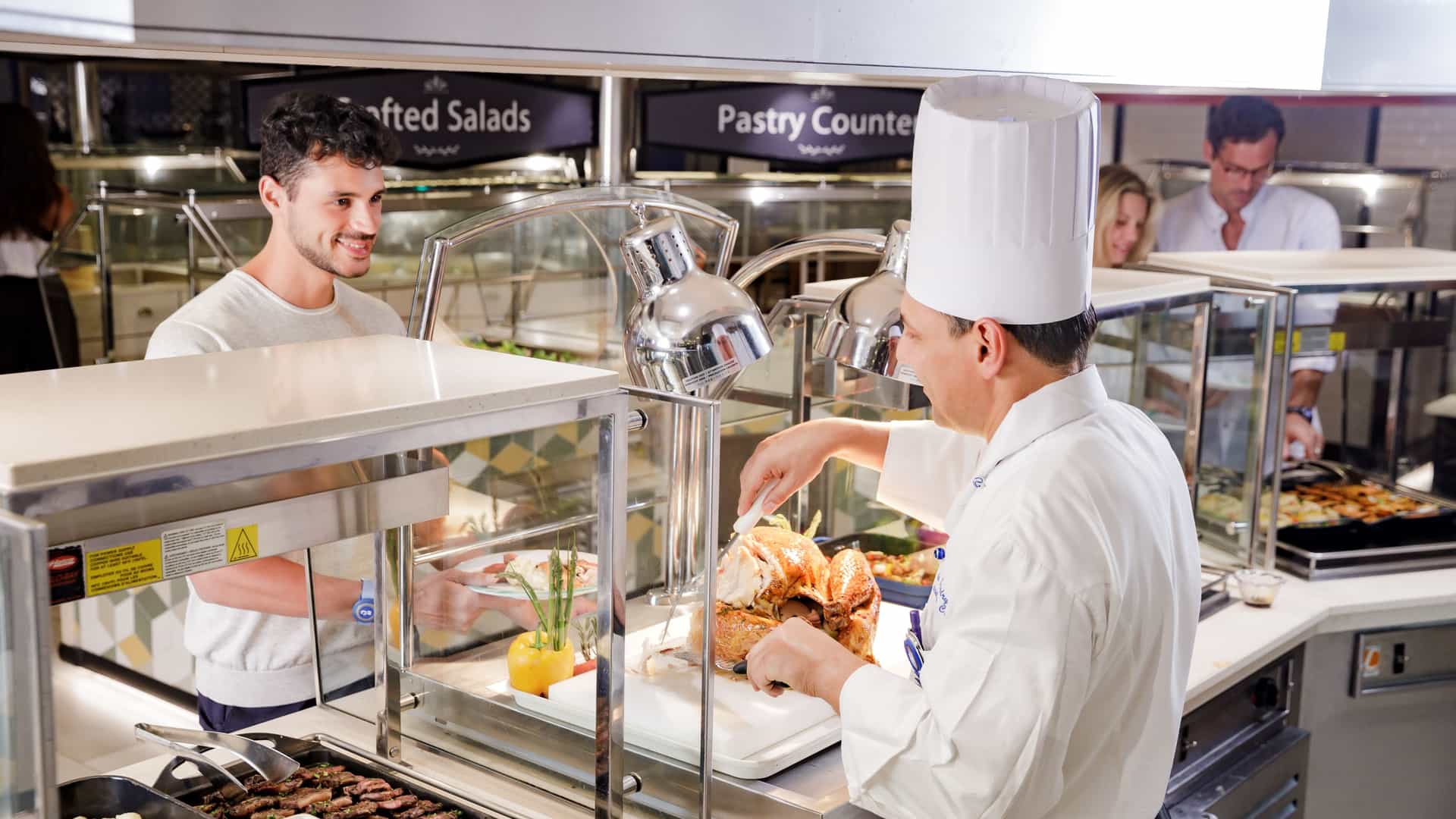 A chef in a white uniform and toque carves roasted meat at a buffet carving station aboard a cruise ship while a guest looks on with a plate.