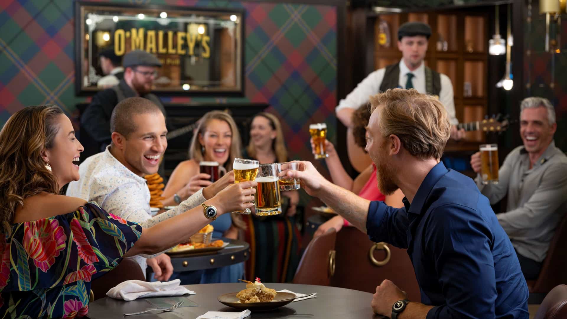 A lively group of patrons raise their glasses for a toast in an Irish pub with plaid decor and a live musician performing in the background.